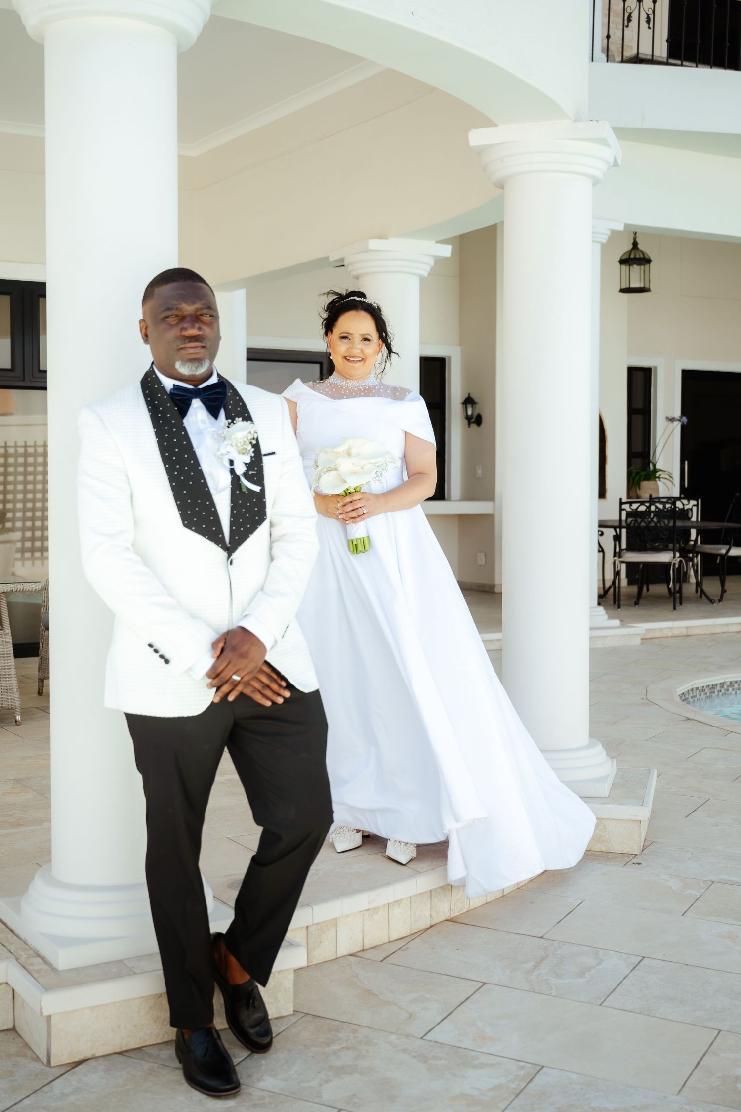 A groom in a white tuxedo and bride in a white gown pose by elegant white columns at their wedding venue.