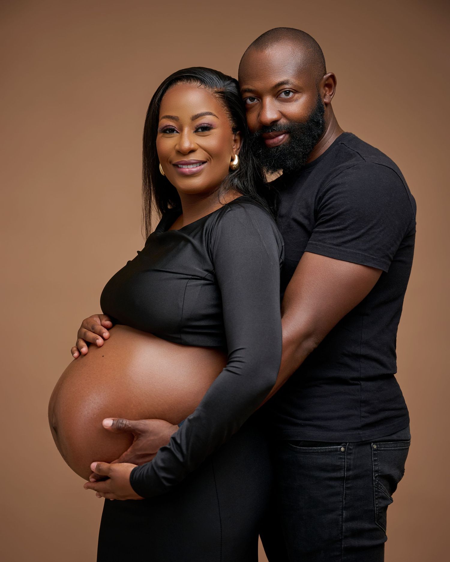 A pregnant woman in a black dress cradles her baby bump while her partner embraces her from behind in a studio maternity shoot.