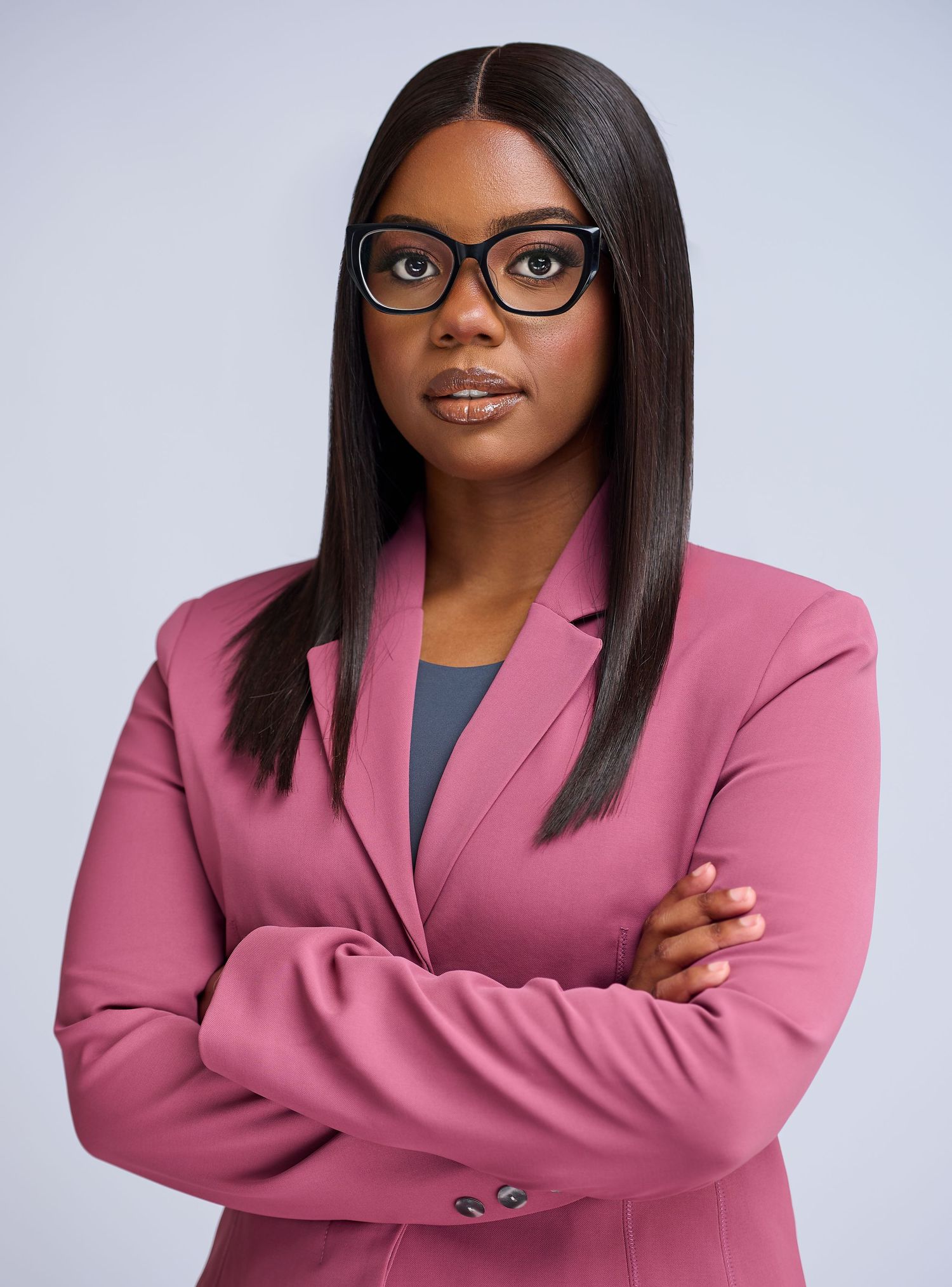 Professional Black woman in pink blazer and glasses stands confidently with arms crossed against gray background.