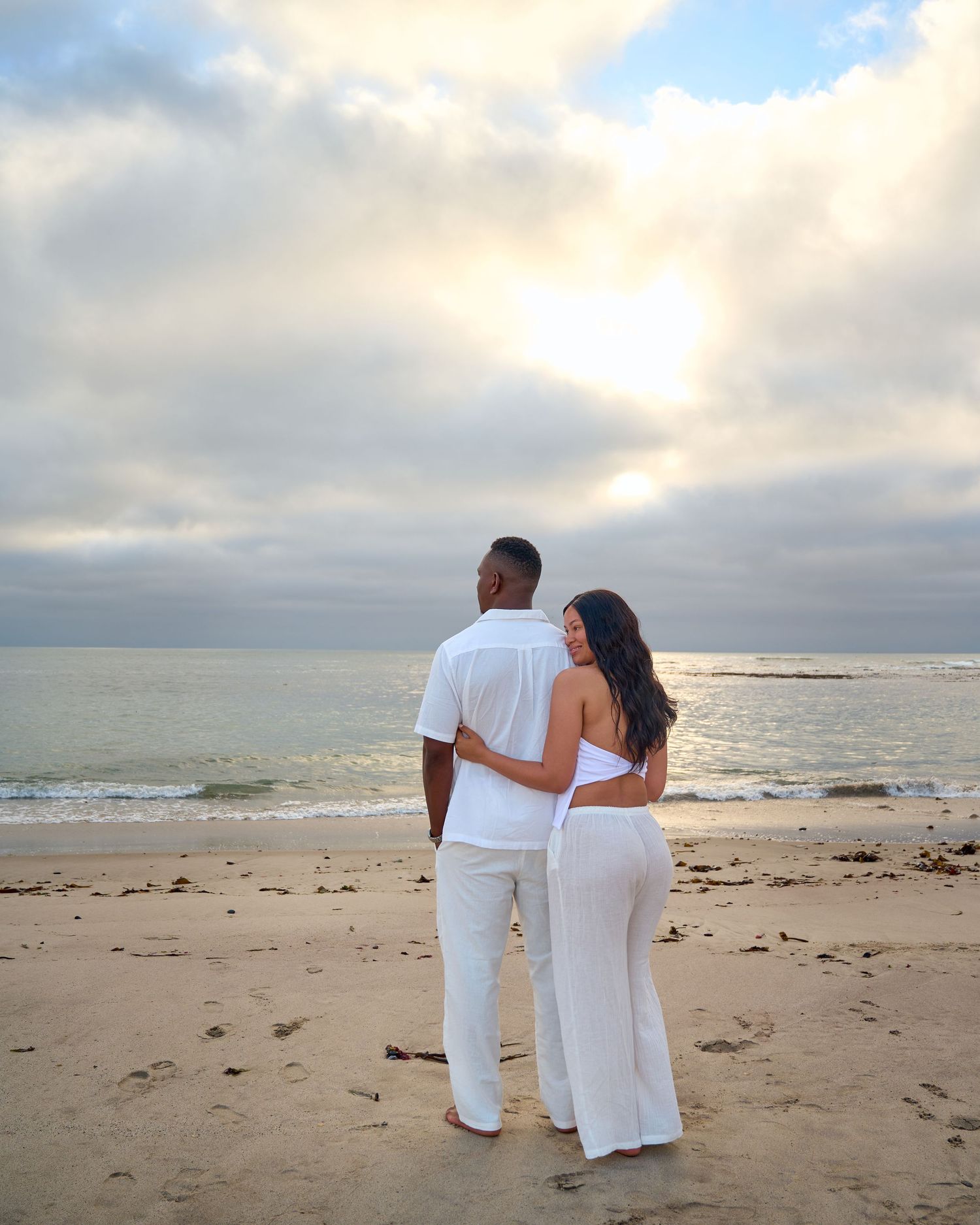Couple in white outfits embracing on a sandy beach at sunset with dramatic cloudy sky and ocean backdrop.