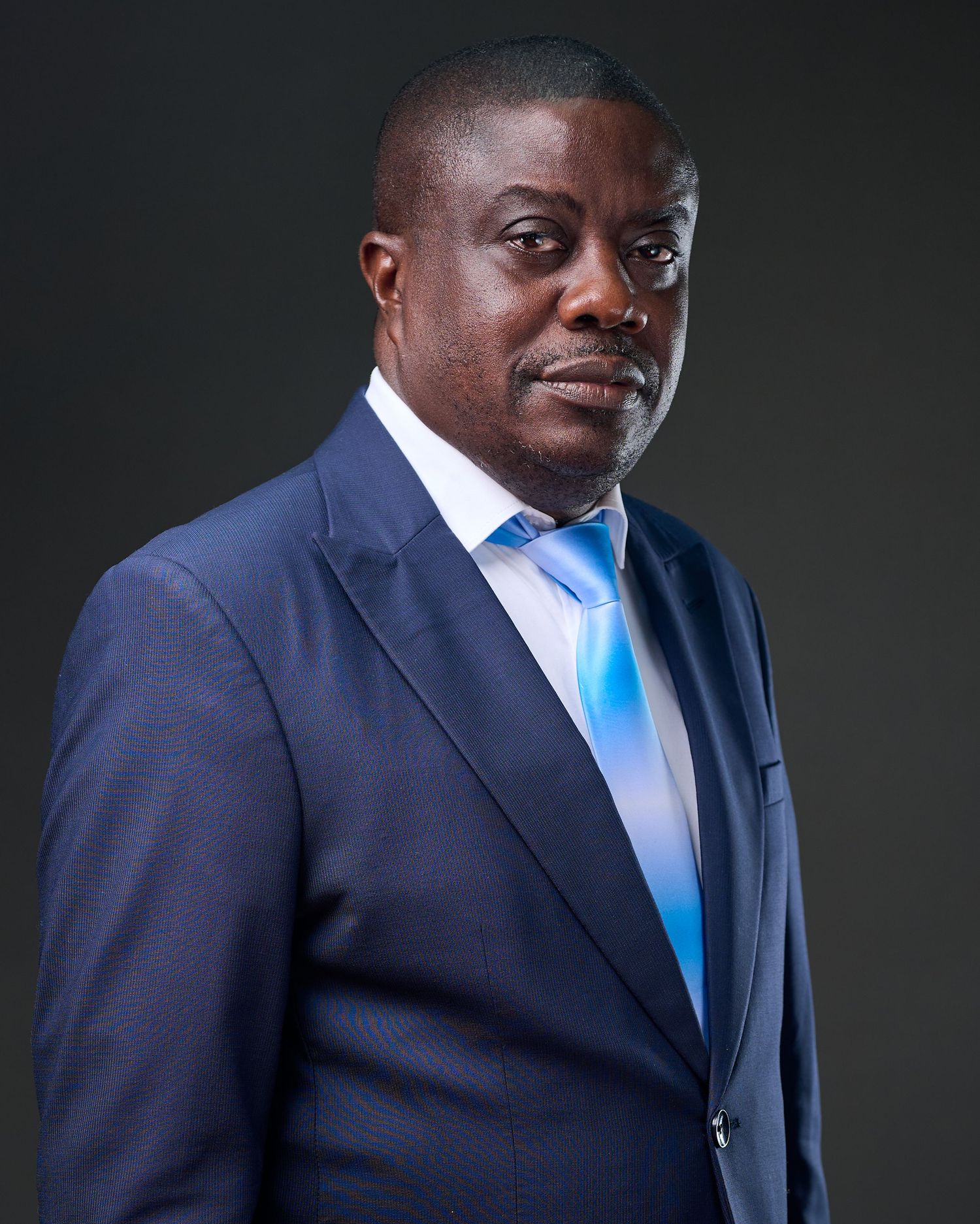 Professional portrait of a person in a navy suit and bright blue tie against a dark background.