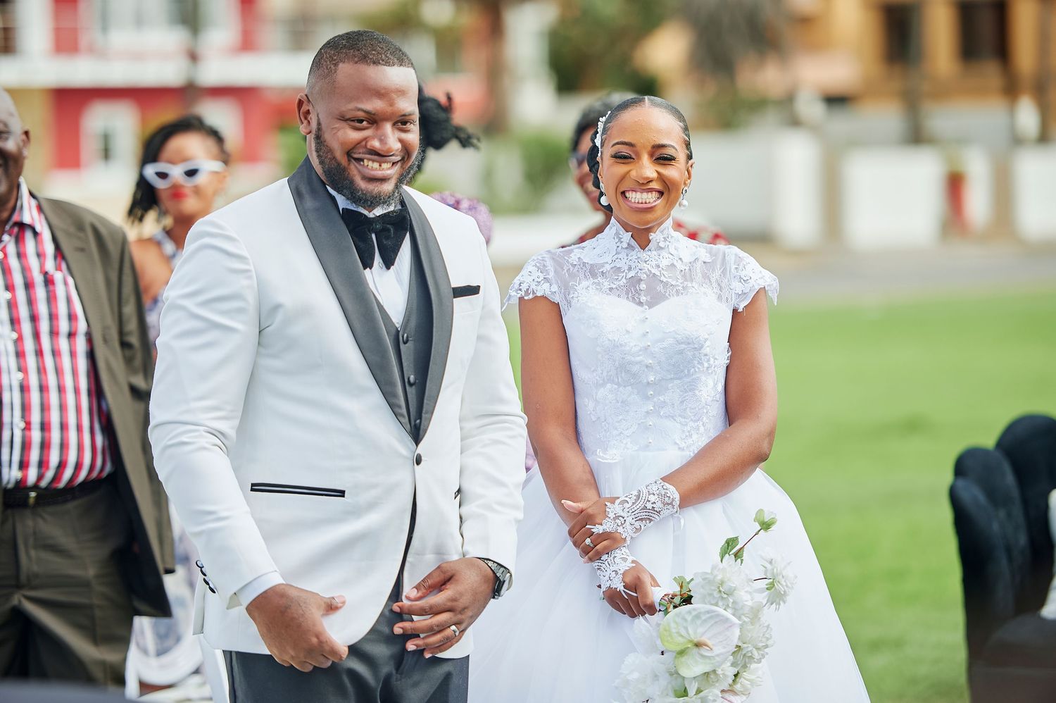 Smiling bride and groom in white wedding attire walking together outdoors during a sunny wedding ceremony.
