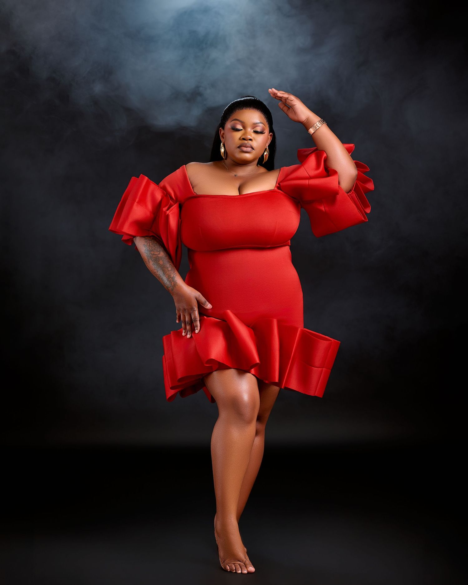 Woman in red ruffled off-shoulder mini dress posing confidently against dark smoky background.