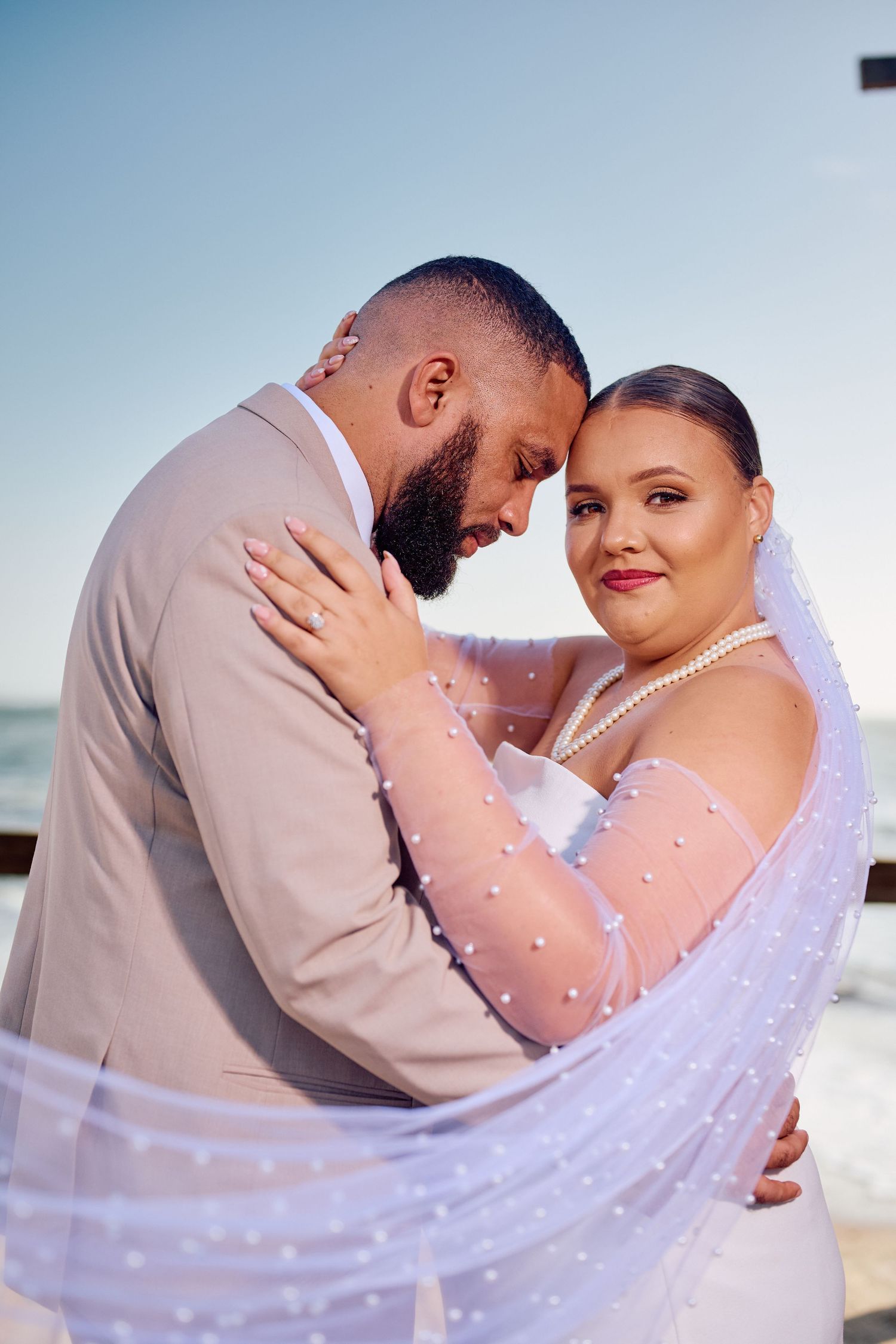 A romantic wedding couple embraces on a beach at sunset in matching white and beige attire.