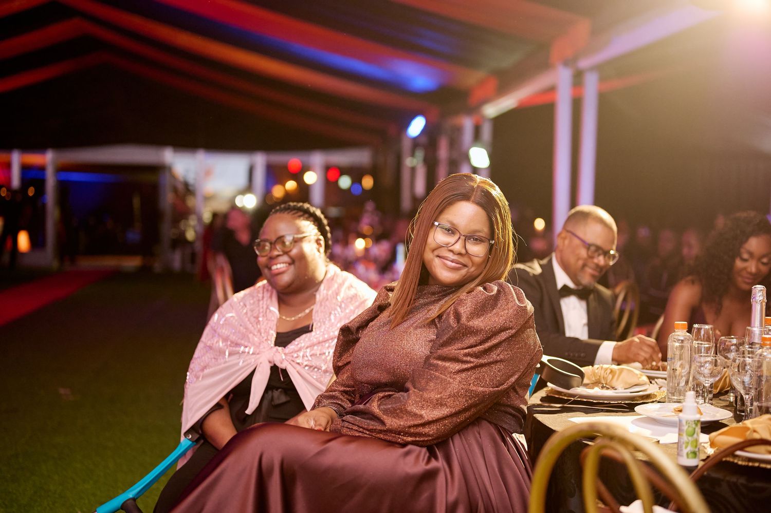 Two women in elegant attire smile at a formal evening gala event with decorated tables and guests in background.