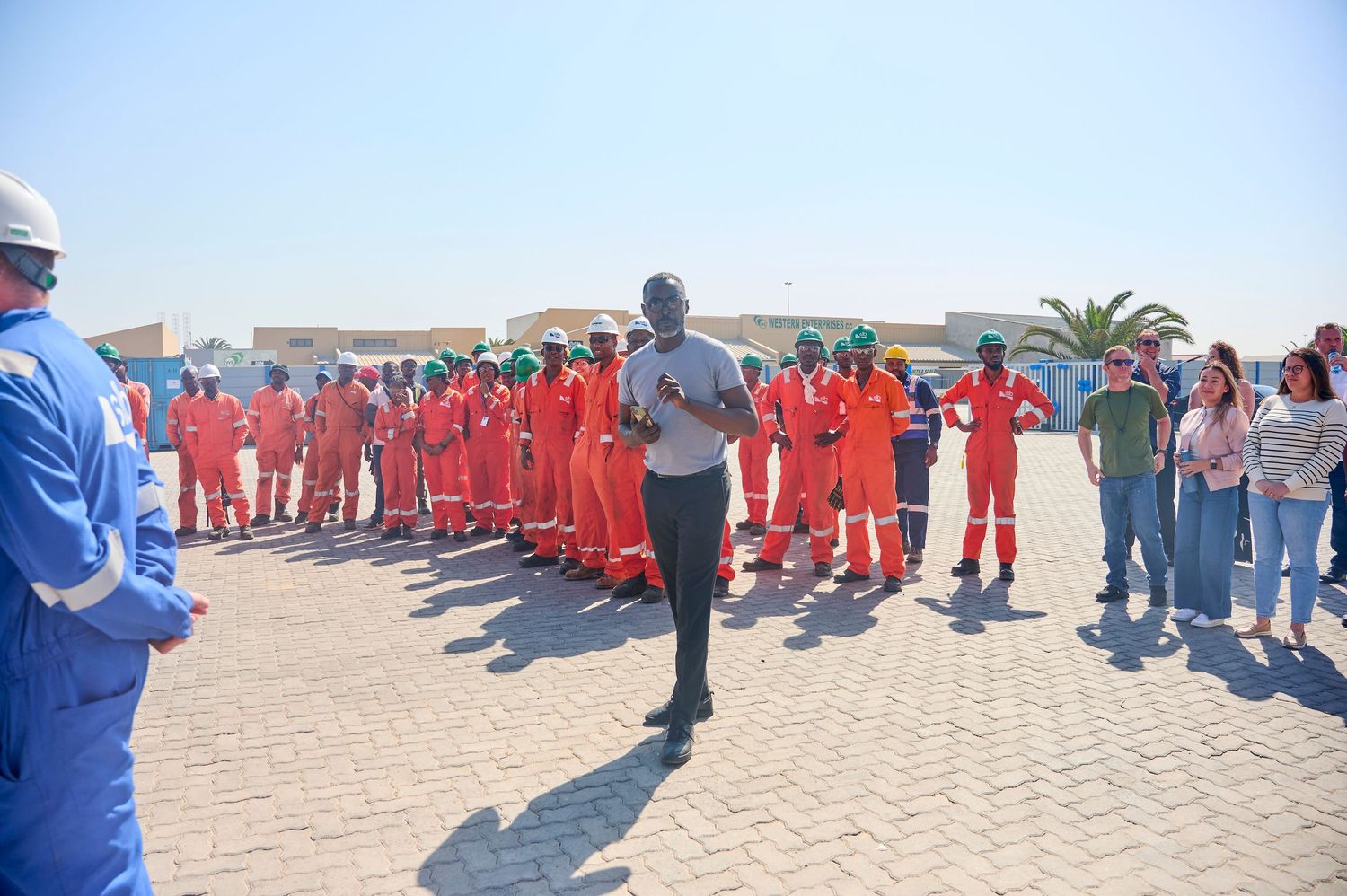 A speaker addresses a large group of workers in orange safety coveralls outdoors at an industrial site.
