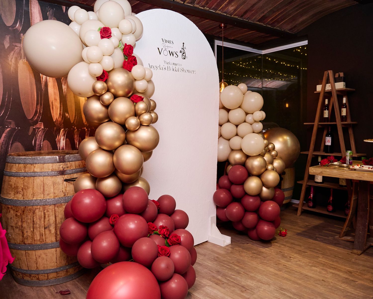 Elegant balloon arch installation with burgundy and gold balloons against a white backdrop near wooden wine barrels.