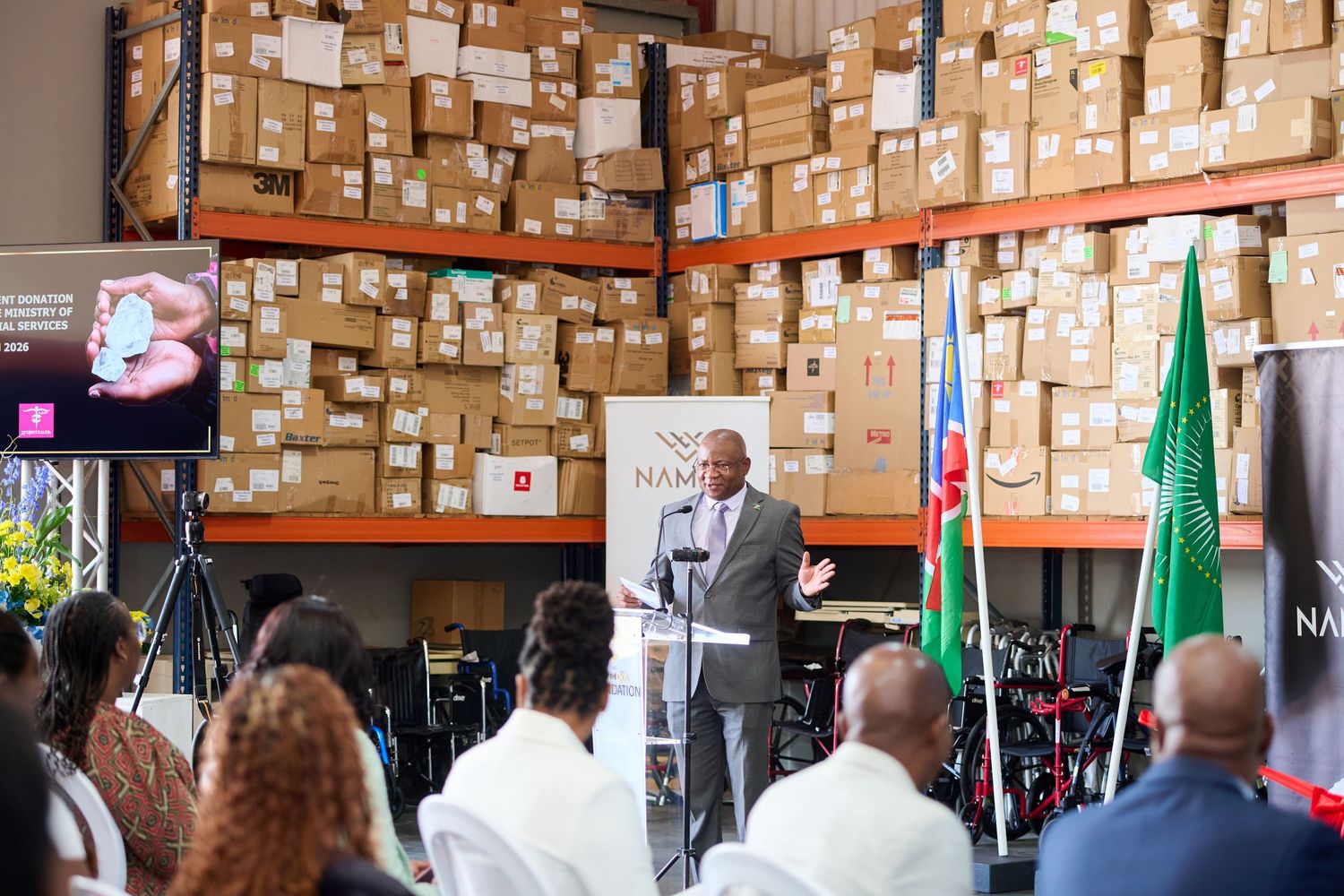 A speaker addresses an audience at a warehouse event with flags and shelves of boxes in the background.