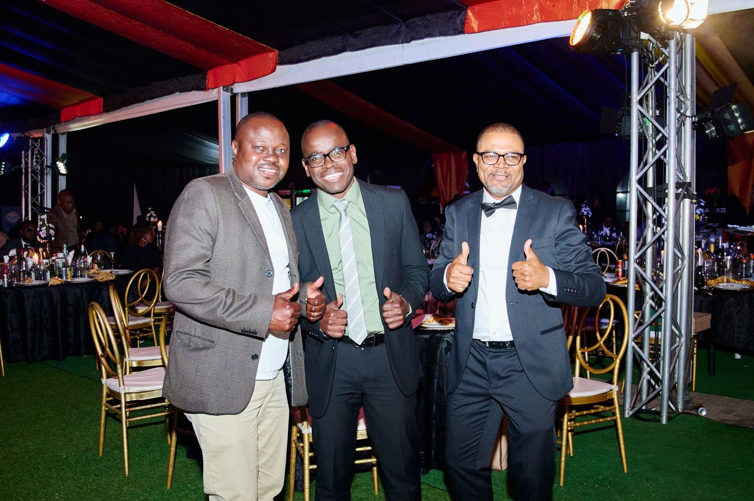 Three men in formal attire smile and give thumbs up at an elegant evening gala event.