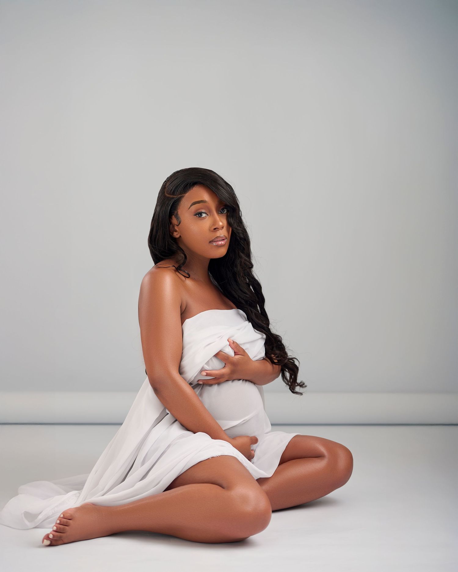 Pregnant woman sitting cross-legged in white draped fabric, cradling baby bump in a professional maternity studio photo.