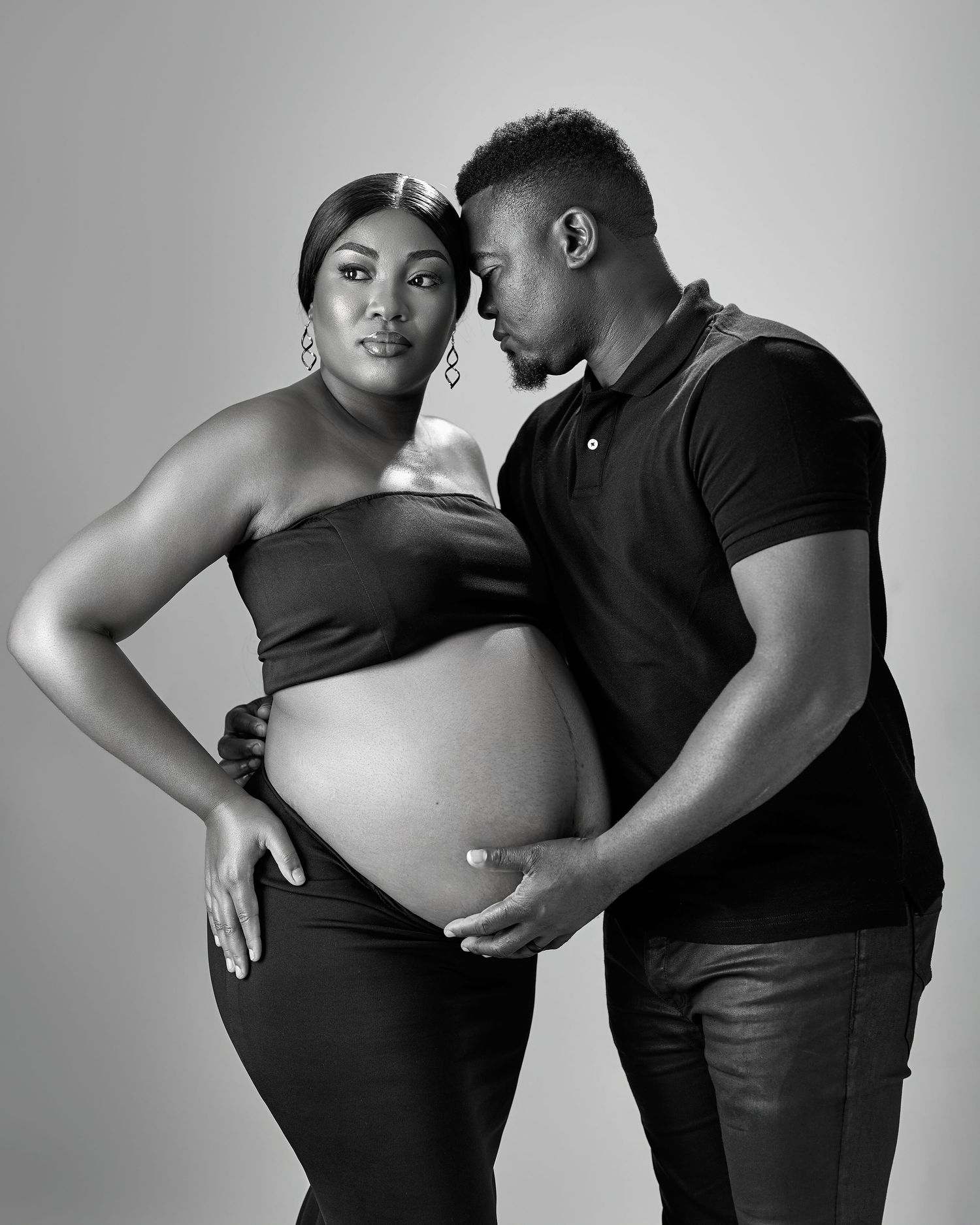 Black and white maternity photo of a pregnant woman and man tenderly posing together in a studio setting.
