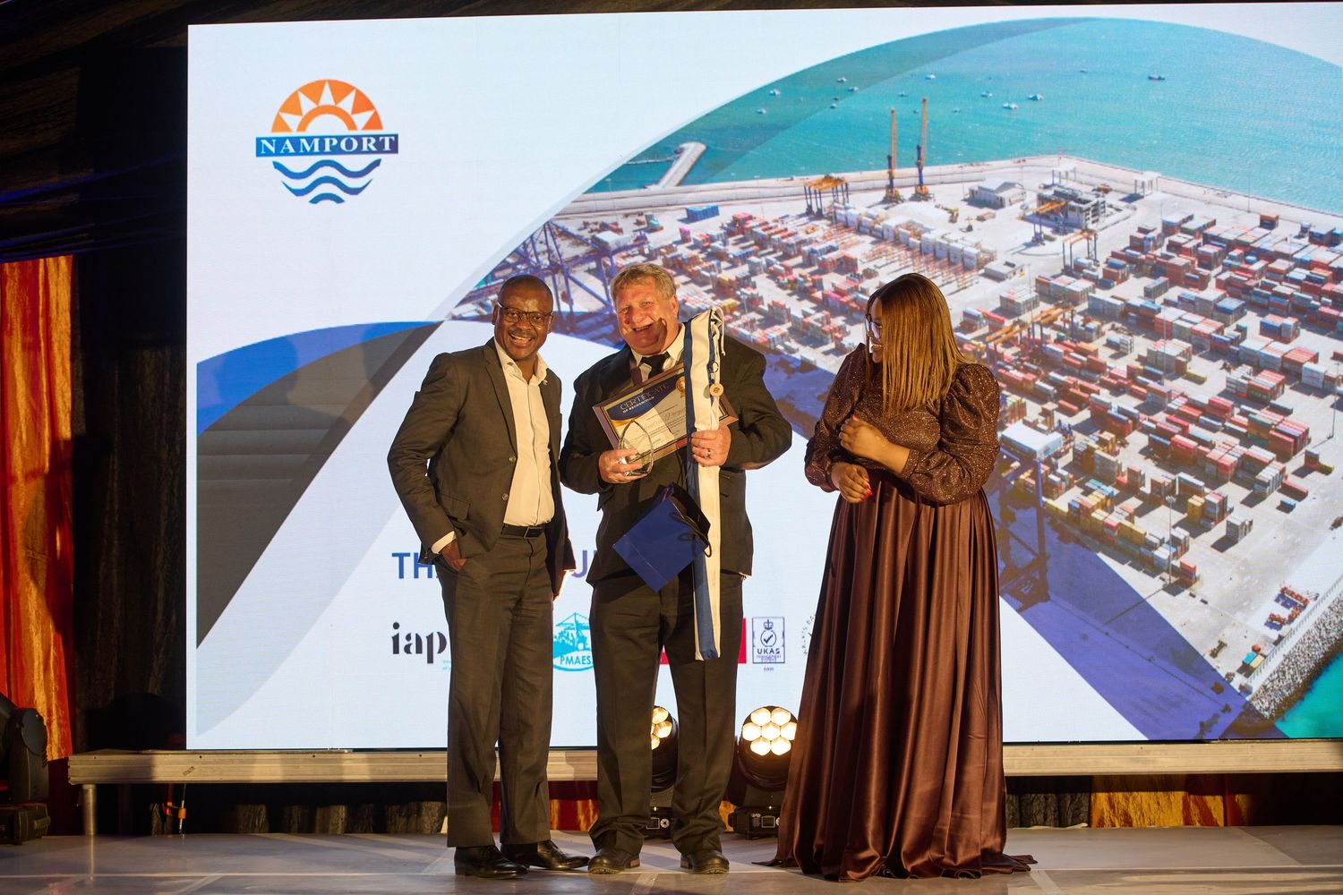 Three professionals pose on stage at a Napier Port awards ceremony with a port aerial view backdrop.
