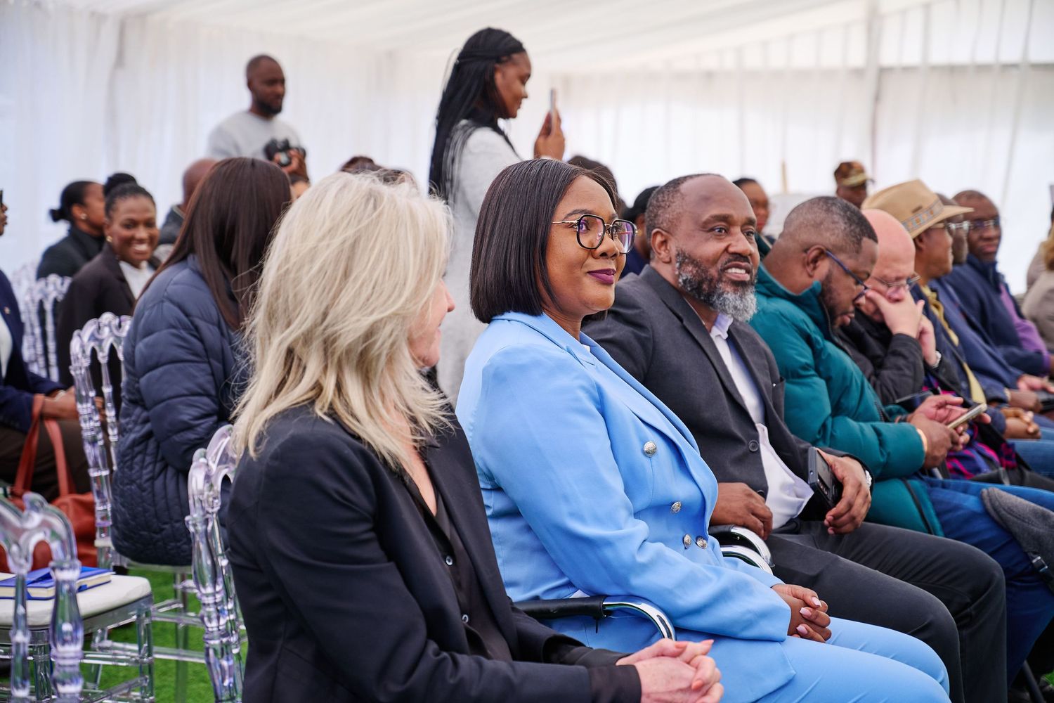 Diverse group of people seated at a formal event under a white tent, smiling and engaged in conversation.