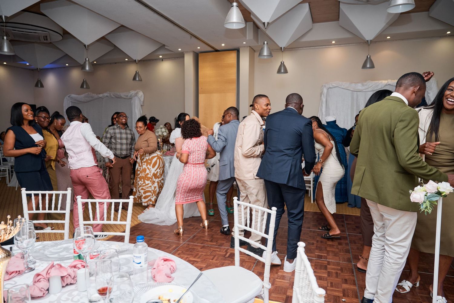 Guests mingling and dancing at an elegant indoor wedding reception with white chairs and pink table settings.