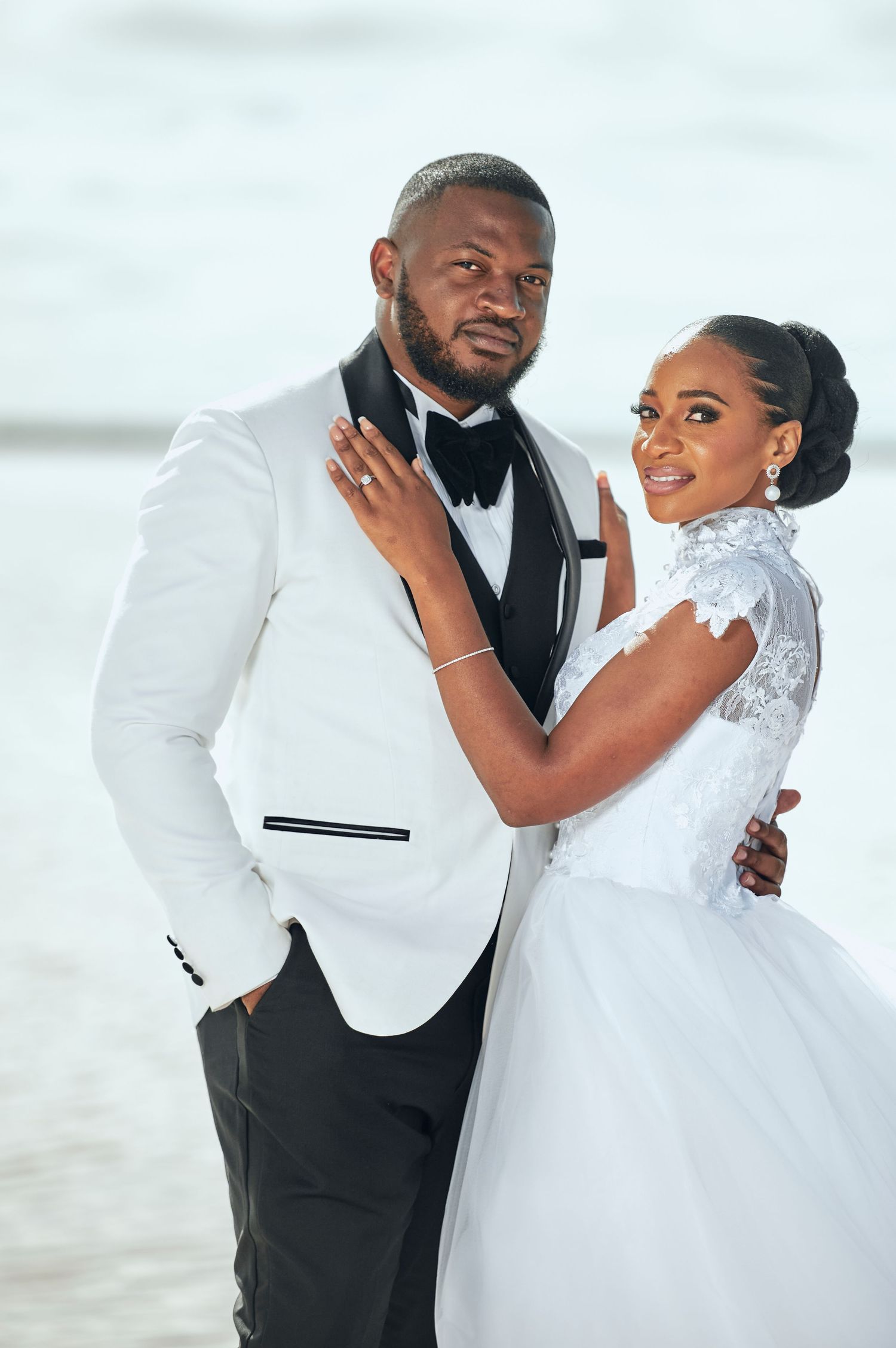 A couple in wedding attire poses on a beach, the groom in a white tuxedo and bride in a white gown.