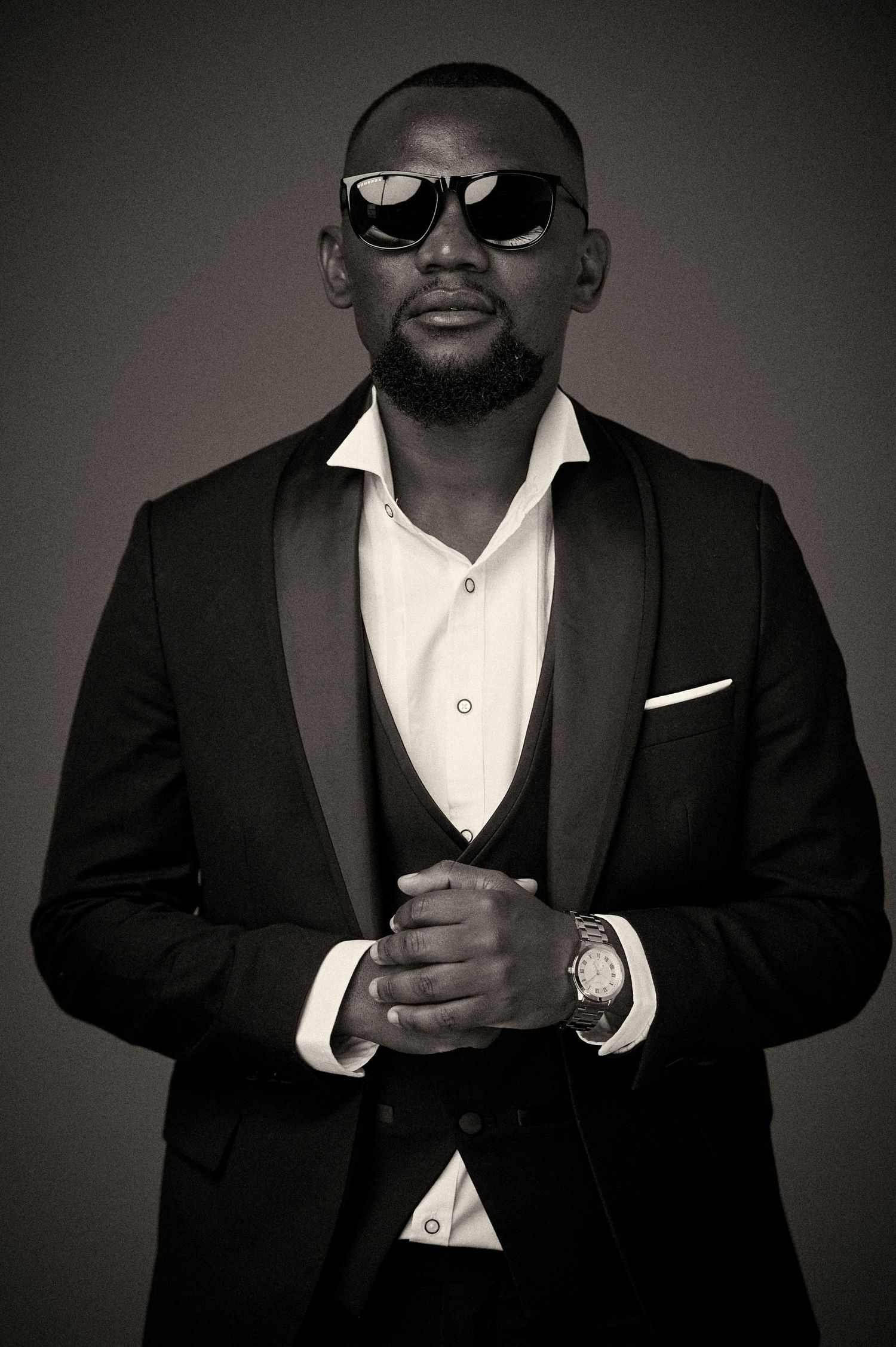 A series of black and white fashion portraits showing a person in formal black suit and sunglasses against a dark background.