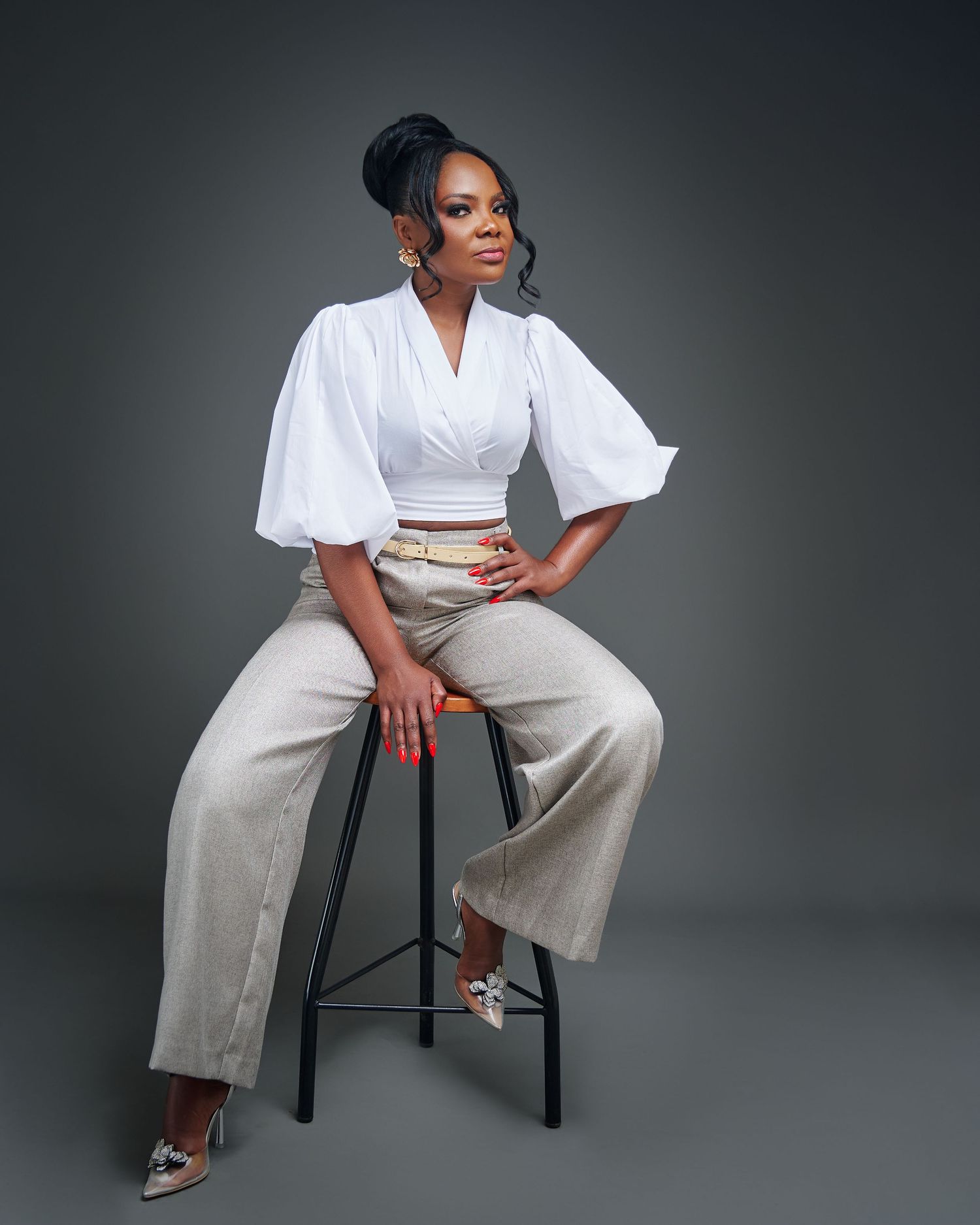 Fashion model in white blouse and beige wide-leg pants poses on black stool against dark studio backdrop.