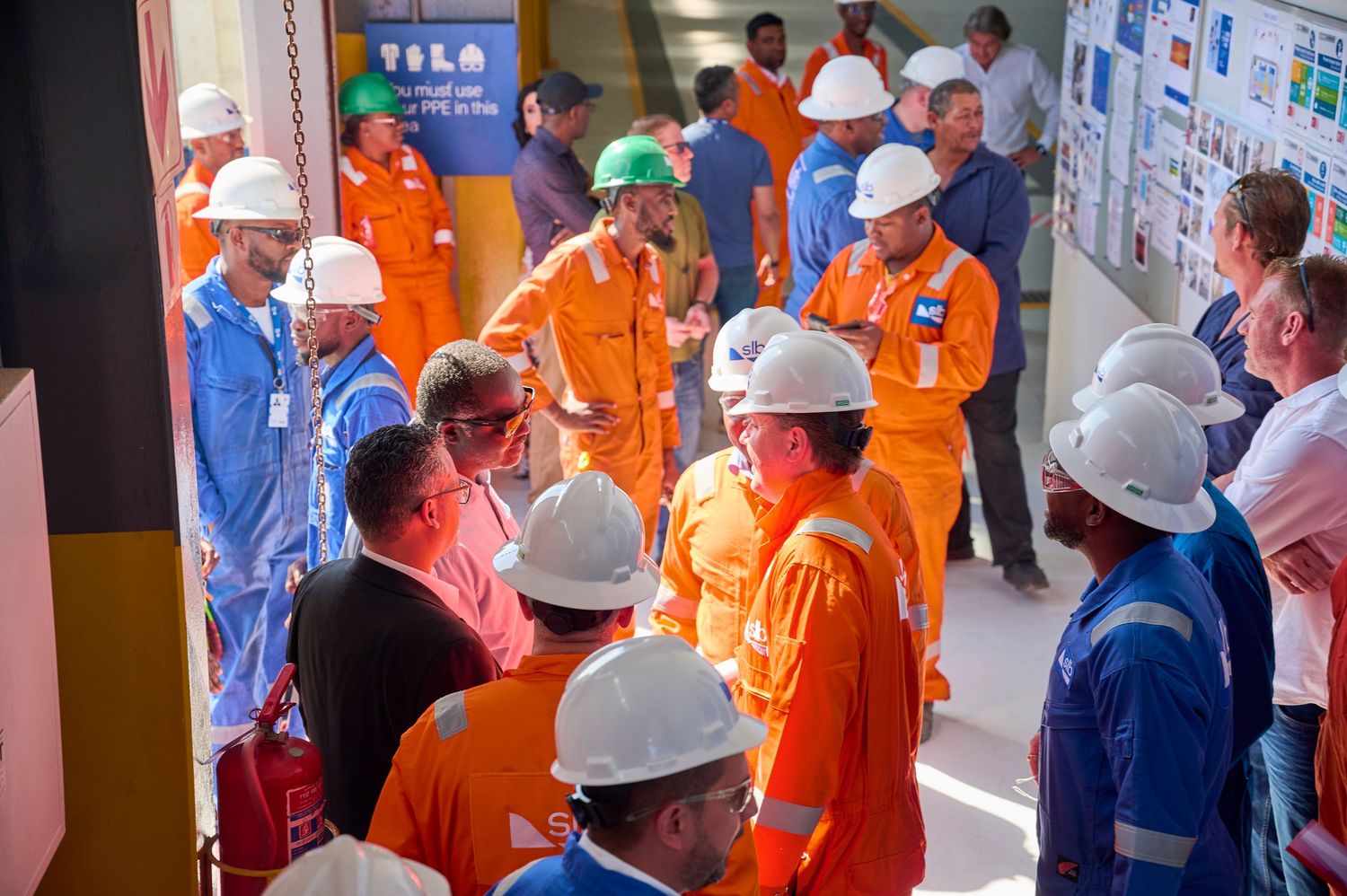 Workers in orange safety gear and white hard hats gather for a safety briefing at an industrial facility.