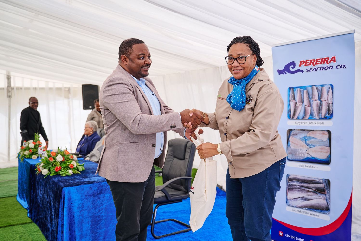 Two people shake hands at a Pereira Seafood Co. event under a white tent with branded signage and fish products displayed.