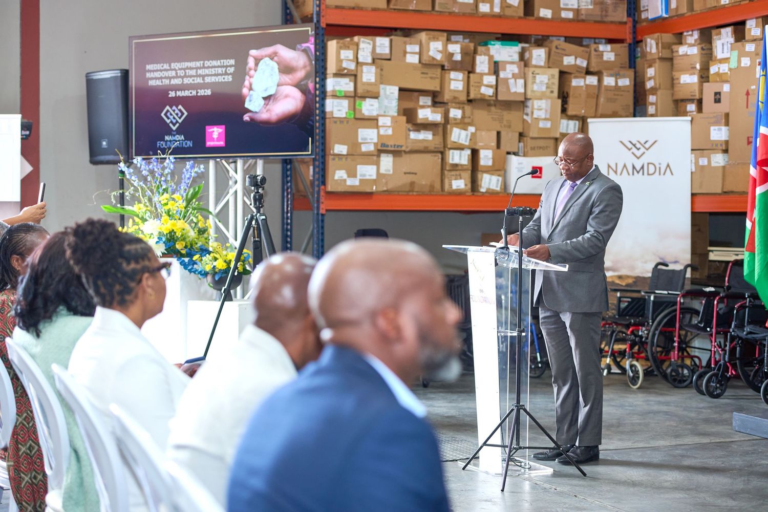 A man speaks at a podium inside a Namdia warehouse during an official event with seated guests and stacked boxes.