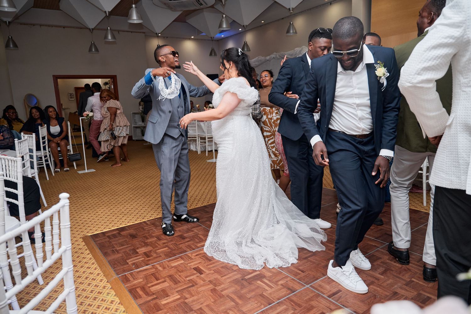 Bride and groom dancing joyfully with guests at a wedding reception on a wooden dance floor.