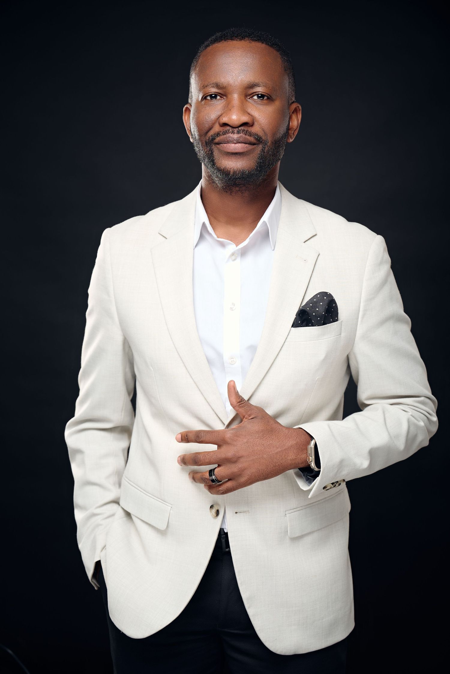 Confident Black man in cream blazer and white shirt poses against dark background with hand on chest.