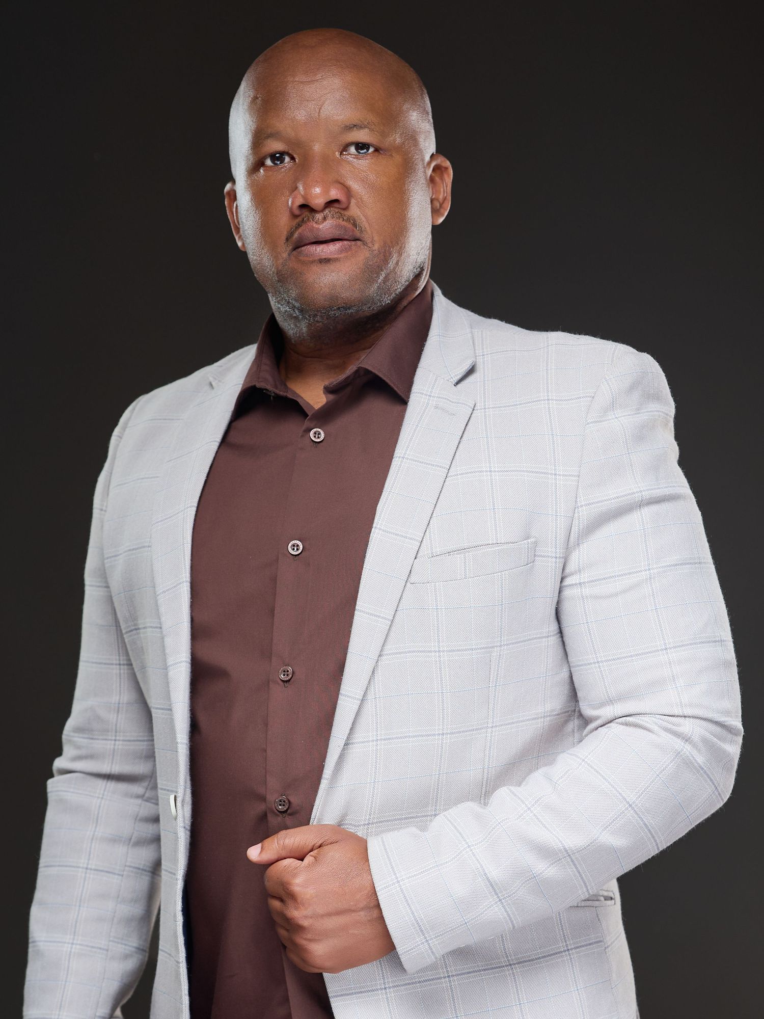 A confident man in a light gray plaid blazer and brown shirt poses against a dark studio background.