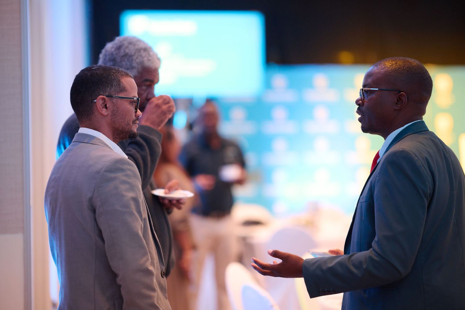 Three business professionals engaged in conversation at a corporate networking event with blue backdrop lighting.