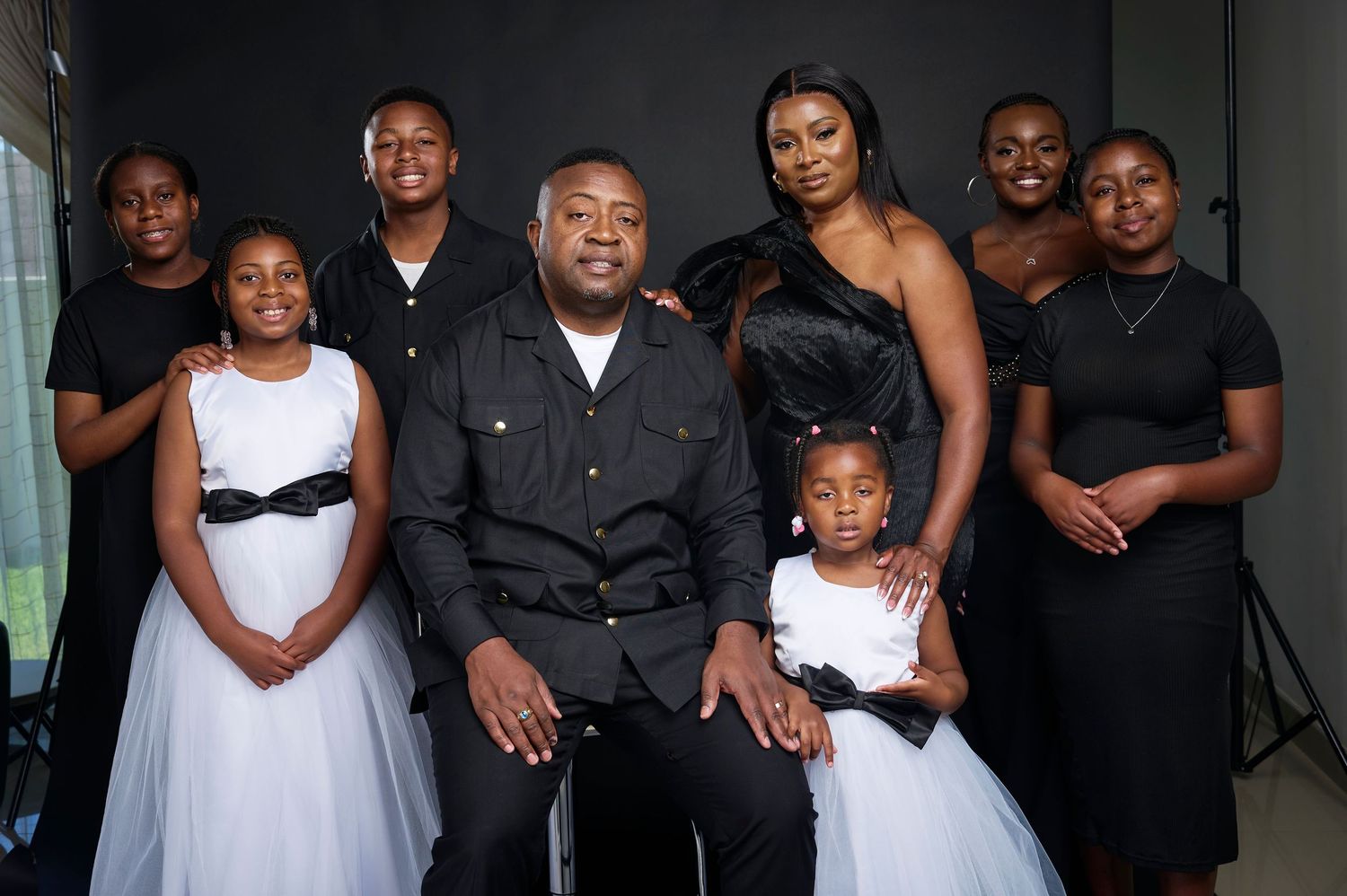 A family portrait of nine people in black and white formal attire posing together against a dark studio background.