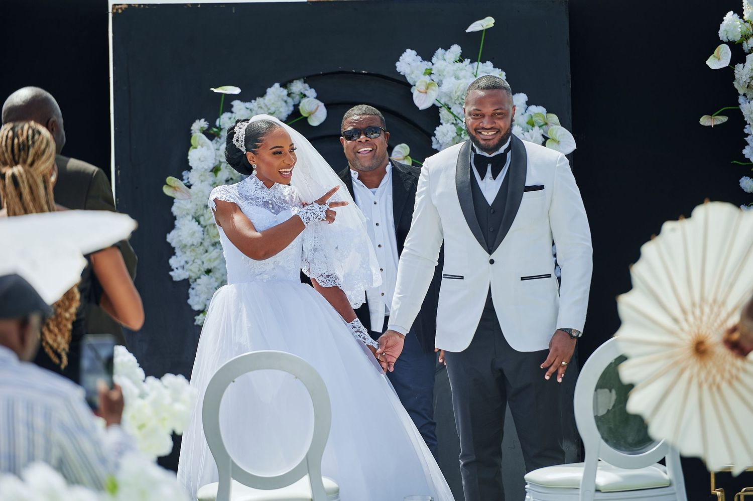 Bride and groom pose with a guest at an elegant outdoor wedding ceremony with white floral decorations.