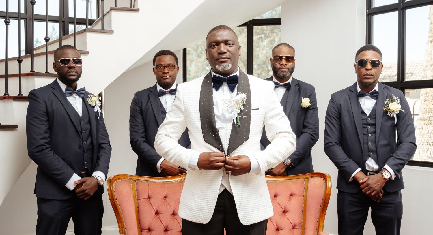Groom in white tuxedo jacket poses with groomsmen in black suits and bow ties at elegant wedding venue.