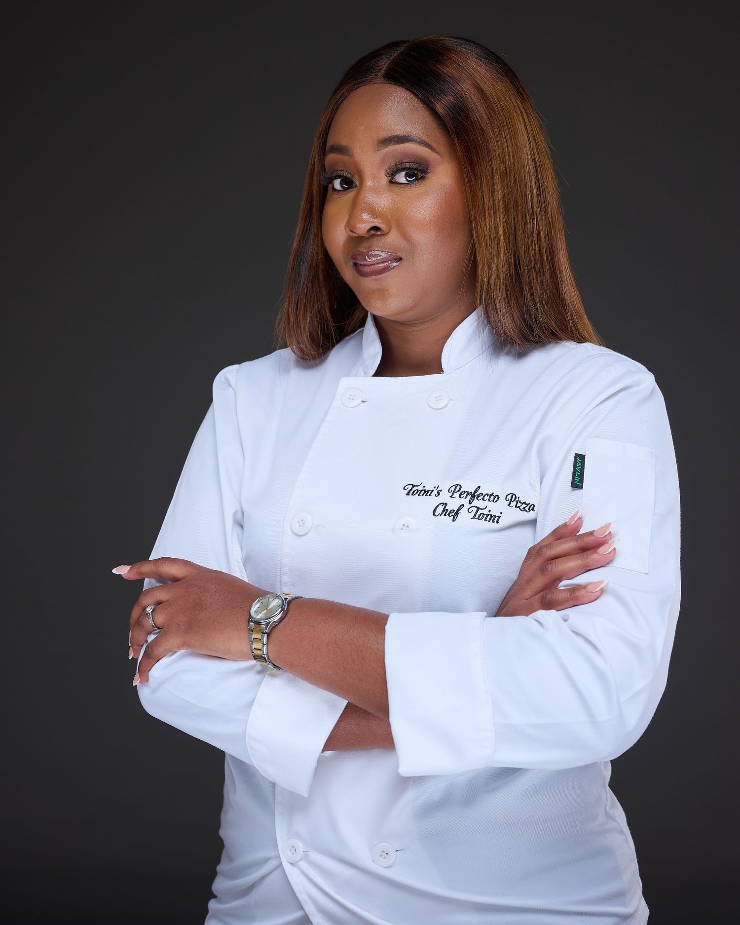 Professional chef in crisp white uniform poses confidently with crossed arms against dark background.