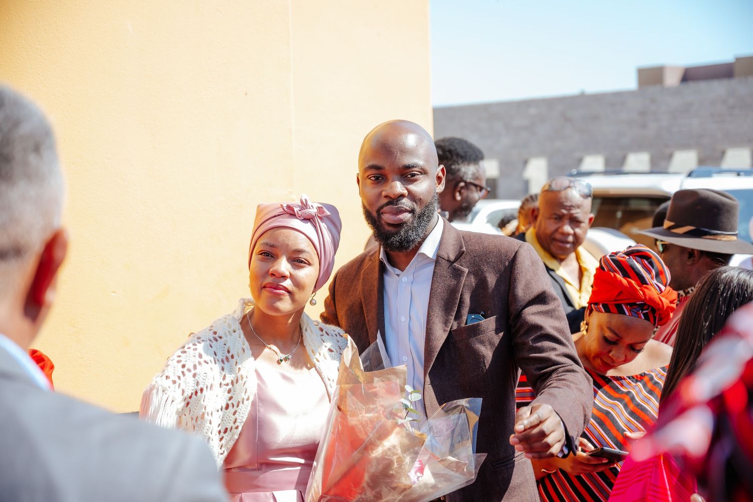 Couple in formal attire smiling at a colorful African cultural celebration outdoors.