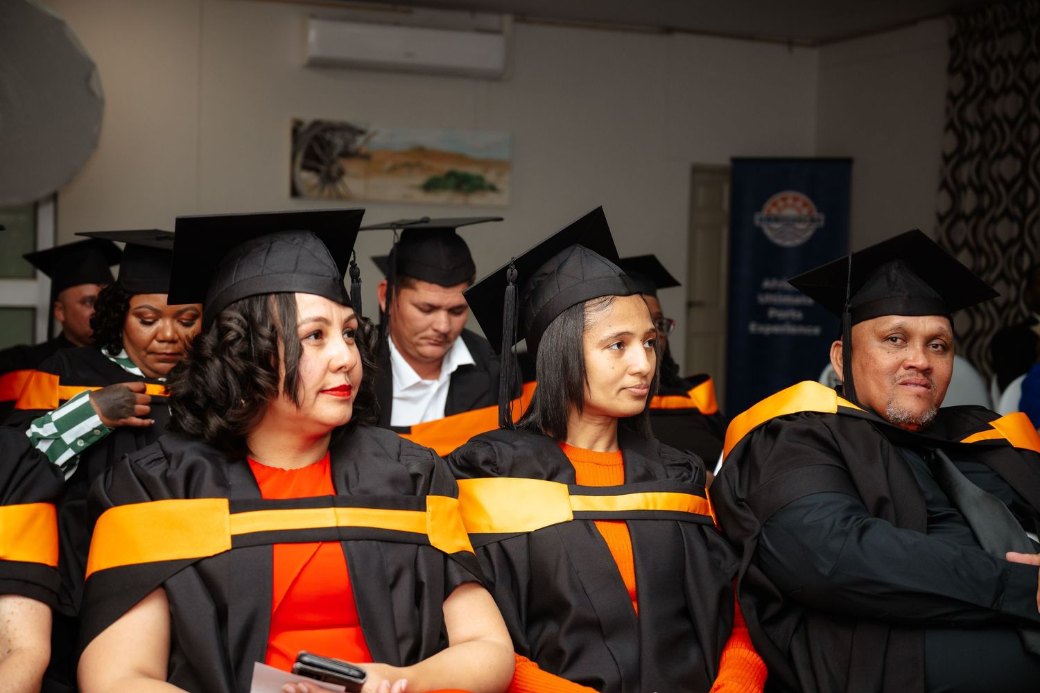 Graduates in black and orange academic robes and caps seated at a graduation ceremony.