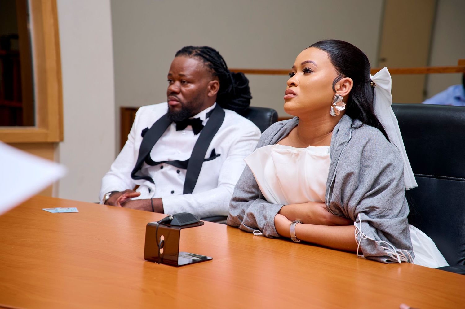 A man in a white tuxedo and a woman in a gray shawl sit together at a wooden conference table.