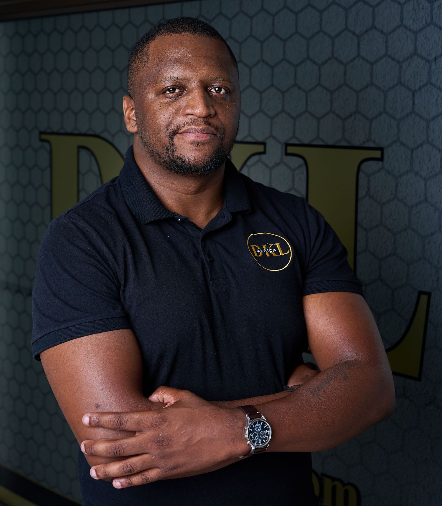 A confident man in a black polo shirt with a logo stands with arms crossed in front of a decorative wall.