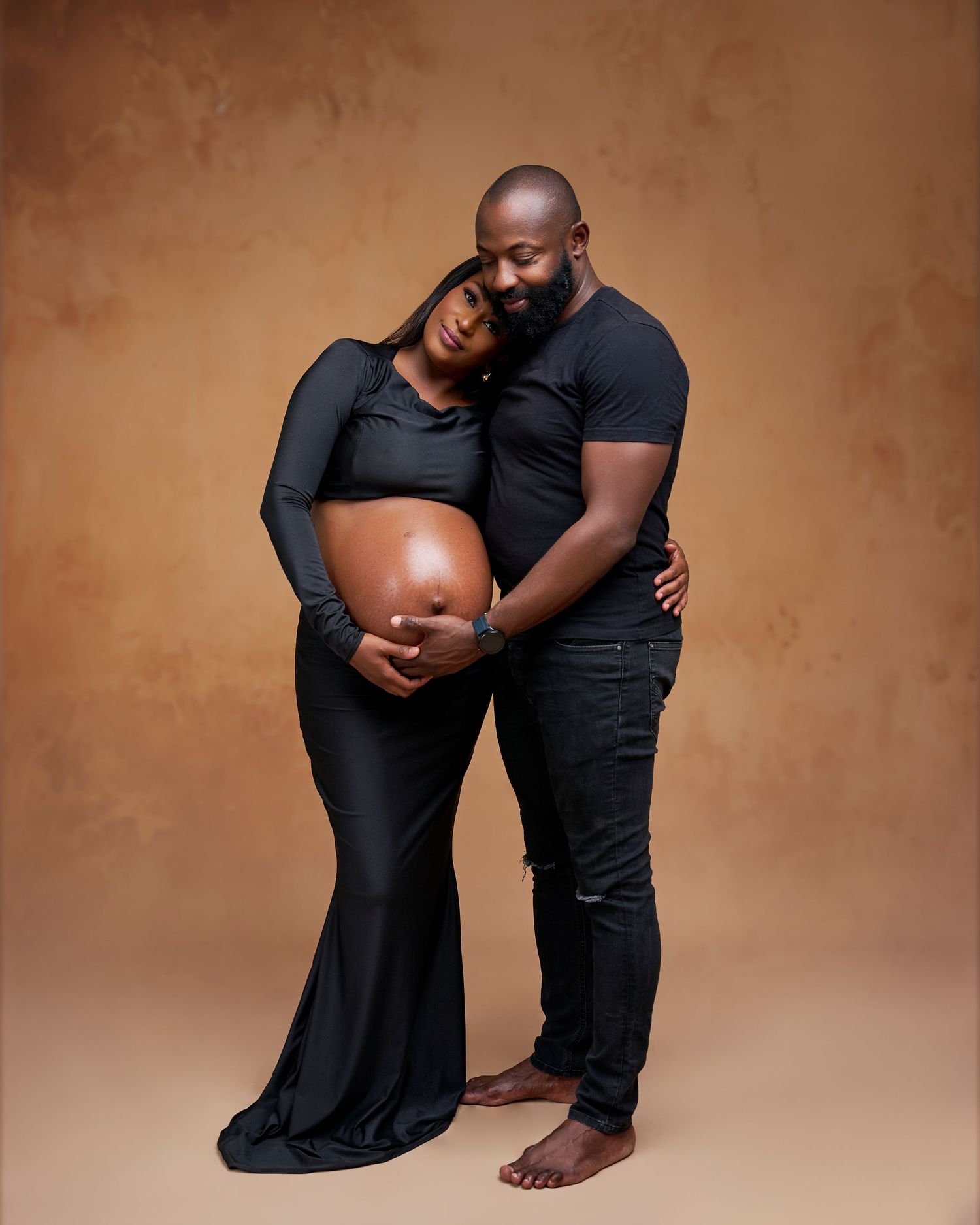 A couple in matching black outfits poses for a maternity photo, the man embracing the pregnant woman from behind.