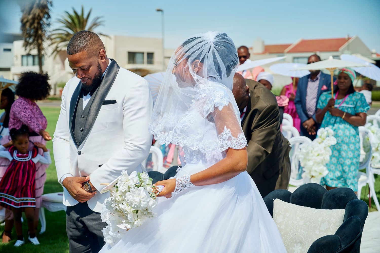 A bride in a white gown and veil walks with a groom in a white suit during an outdoor wedding ceremony.