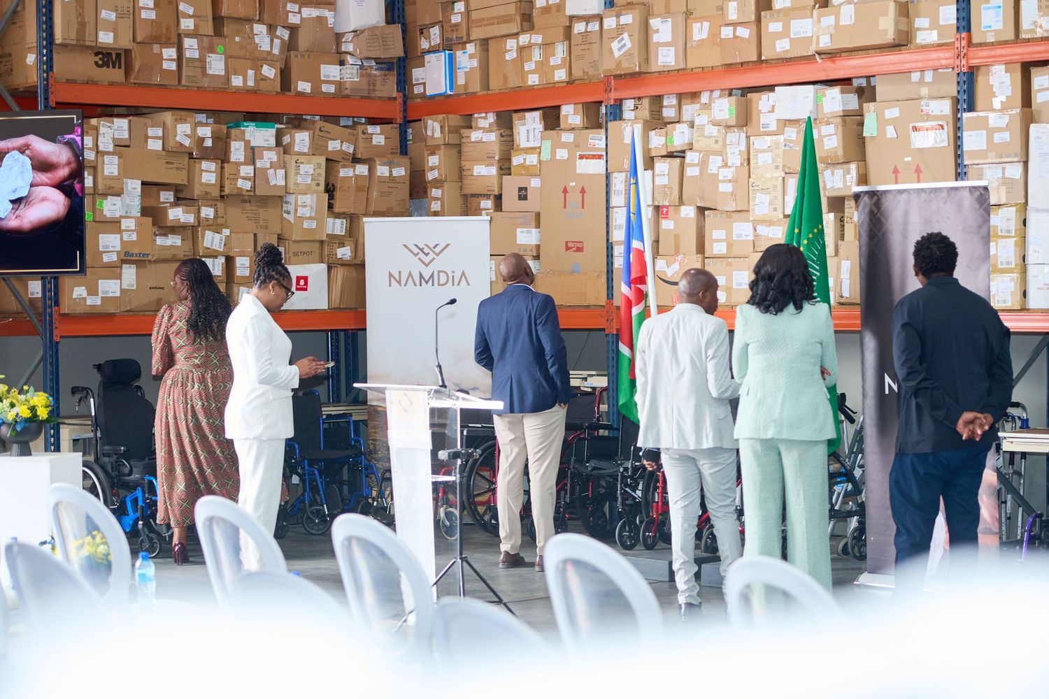 Group of people gathered at a NAMDIA warehouse event with flags and boxes stacked on shelves in background.