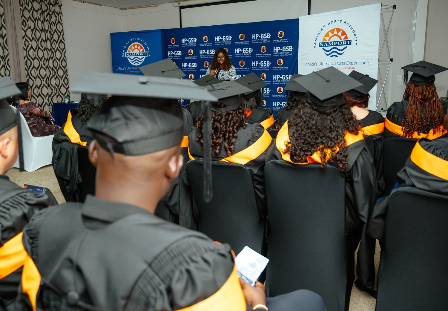 Graduates in black and gold robes attend a graduation ceremony with a speaker at the front.
