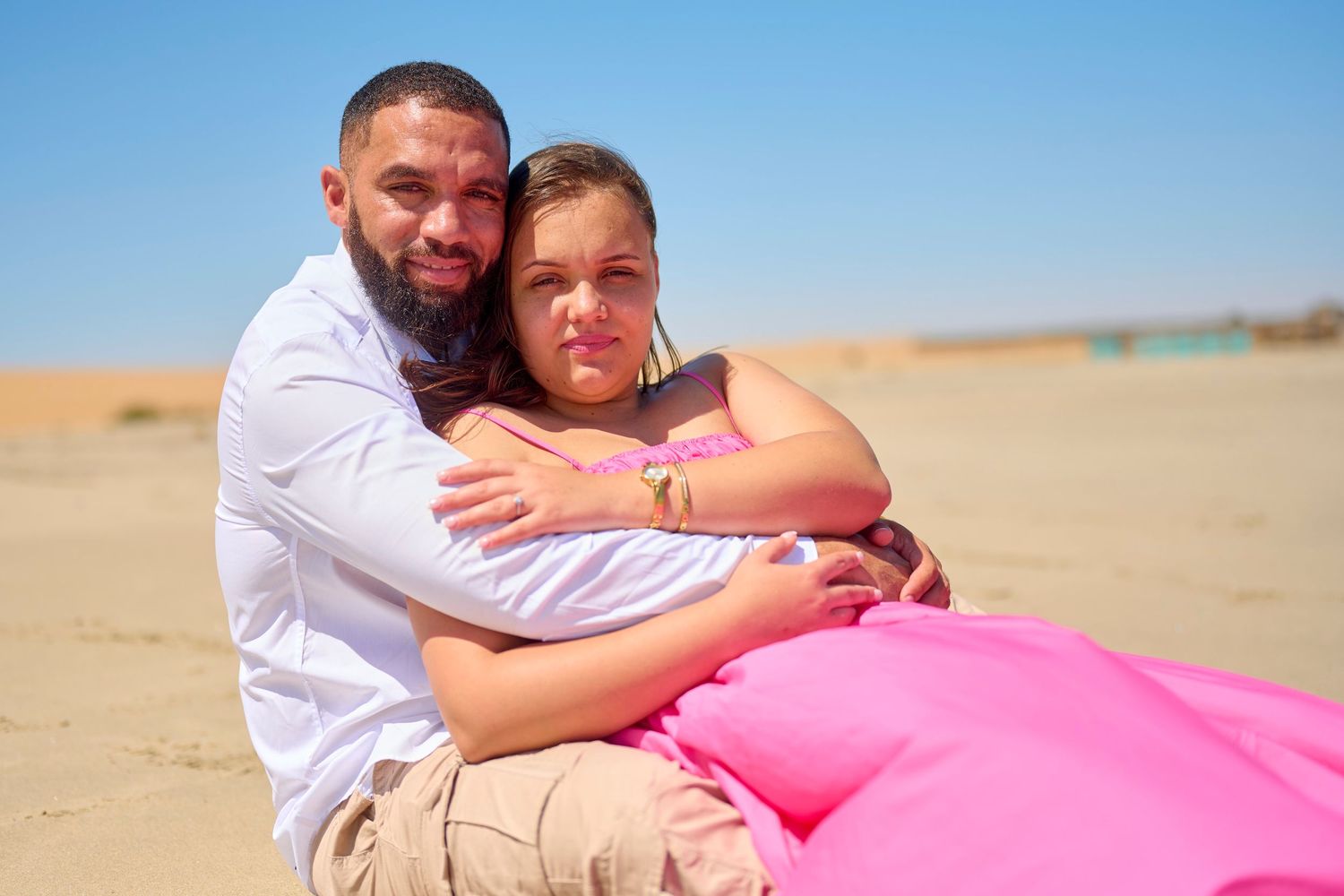 A man and woman share a warm embrace on a sandy beach under a clear blue sky, she wearing a pink outfit.