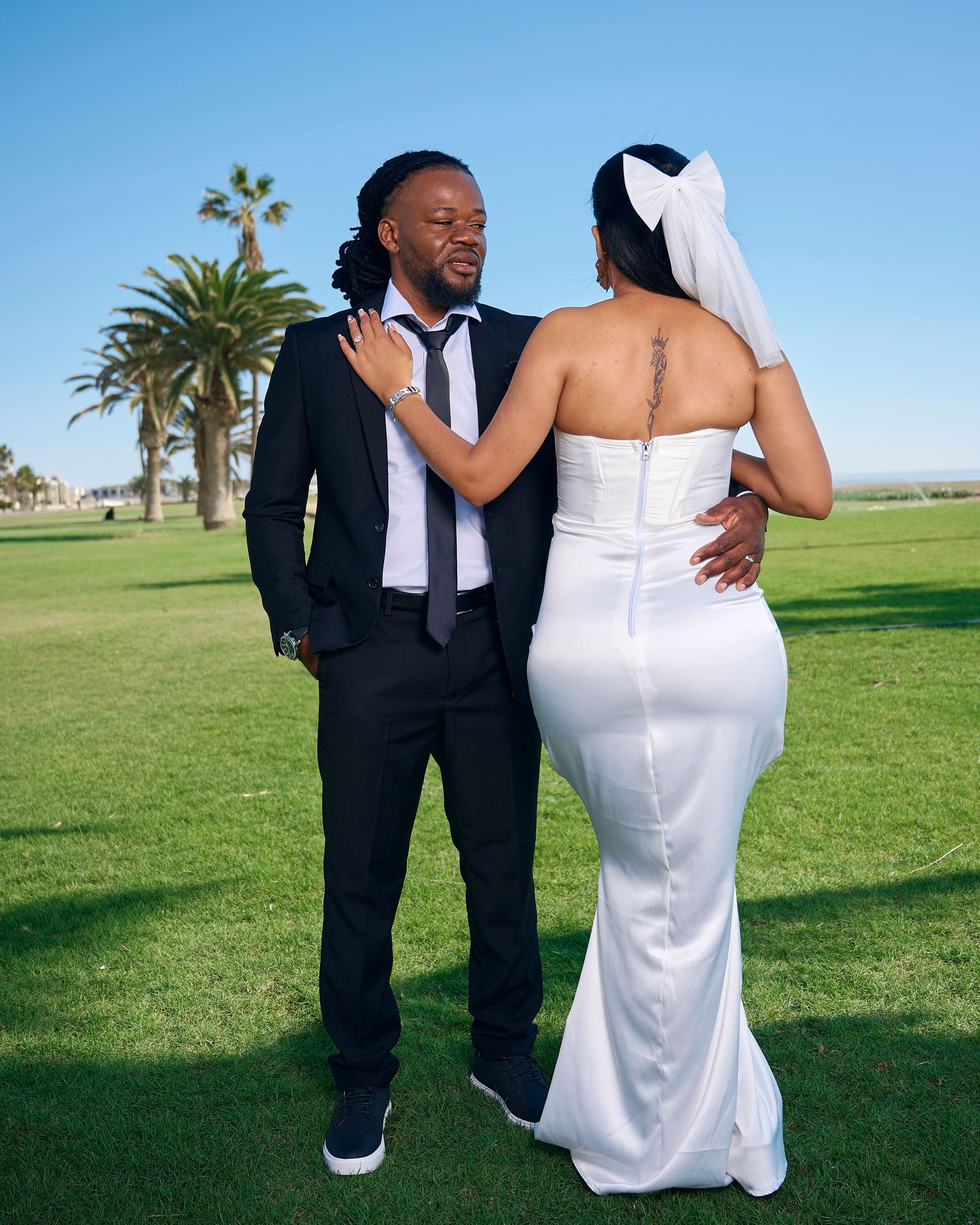 A couple in wedding attire poses on green grass, man in black suit, woman in white mermaid gown with veil.