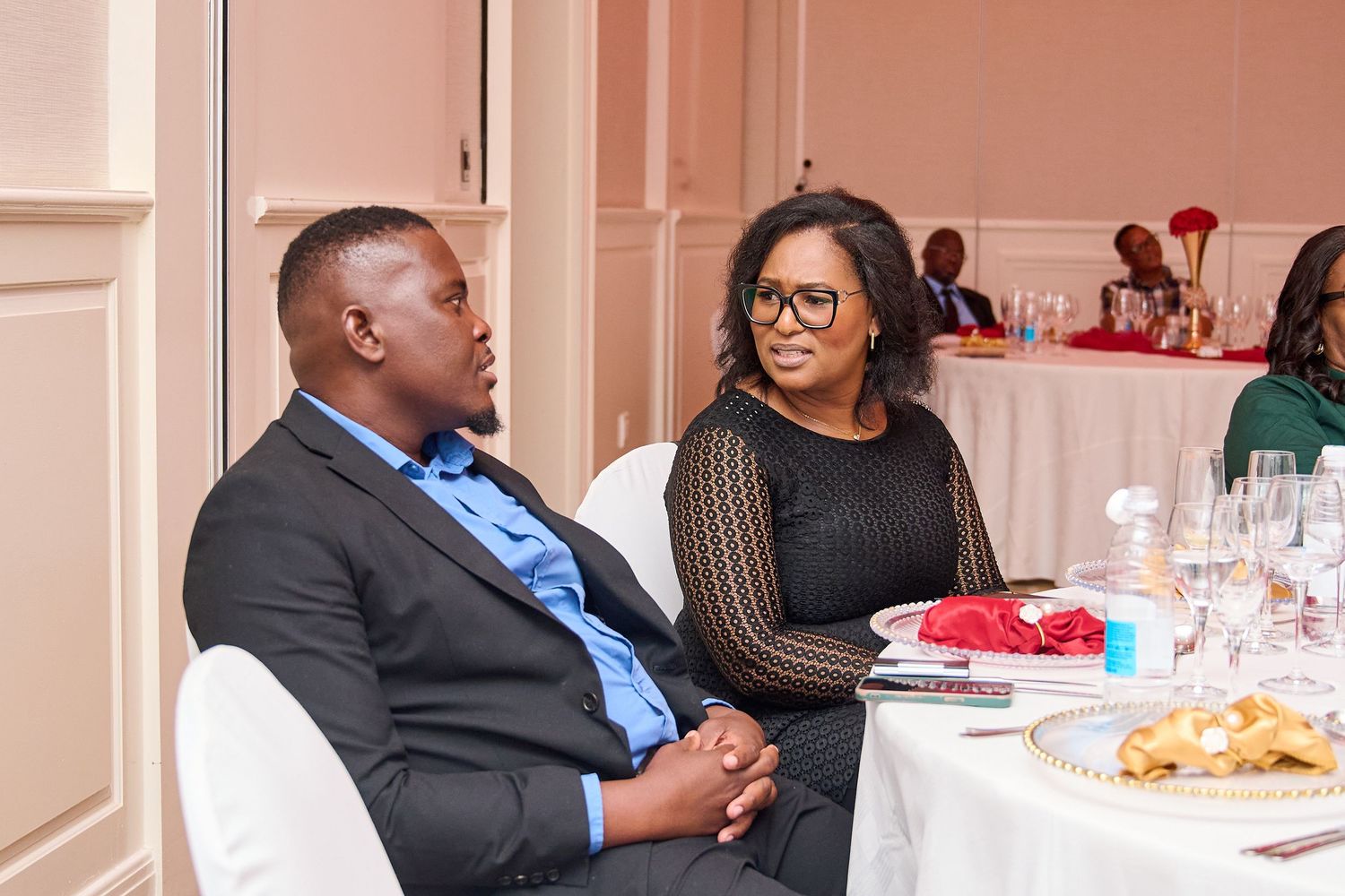 Man in suit and woman in black dress sitting at a elegantly set dinner table engaged in conversation.