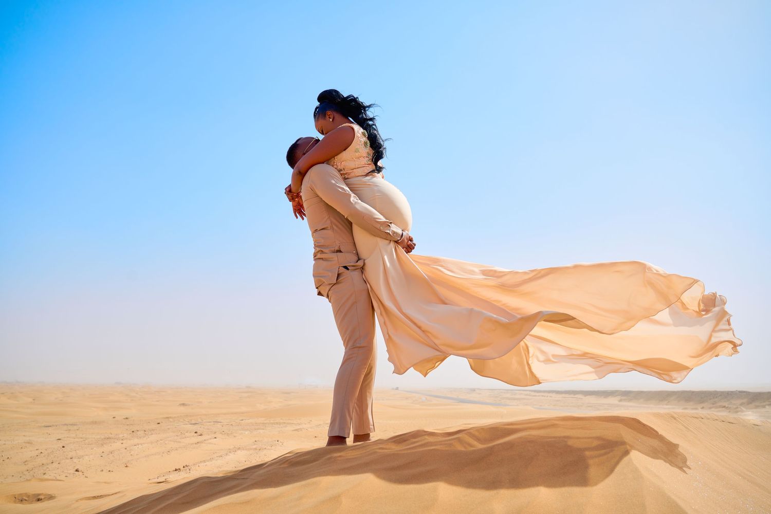 Romantic couple embracing on golden sand dunes in the desert, woman's flowing peach dress billowing in the wind.