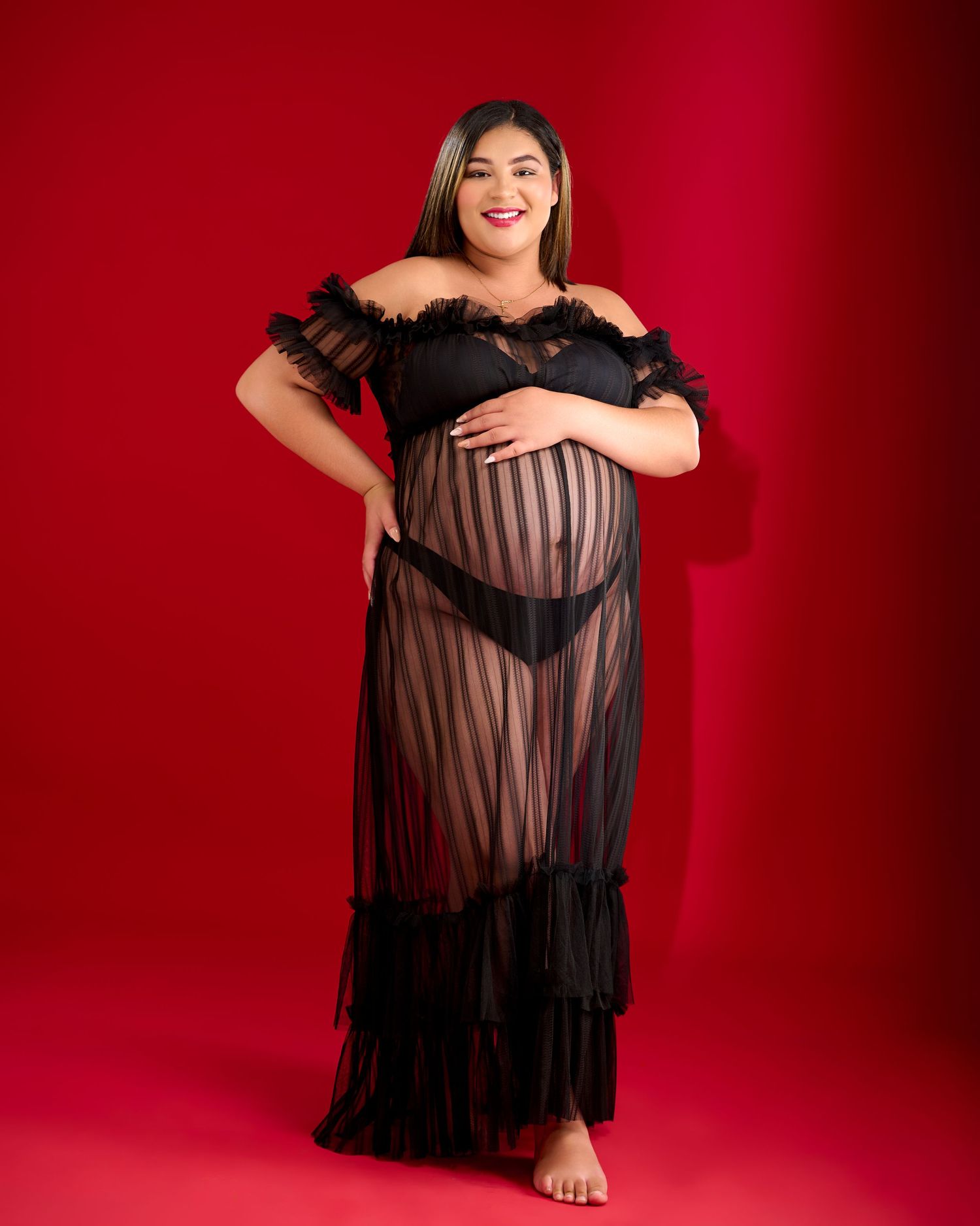 A person in a black lace off-shoulder gown with sheer striped details poses against a red backdrop.