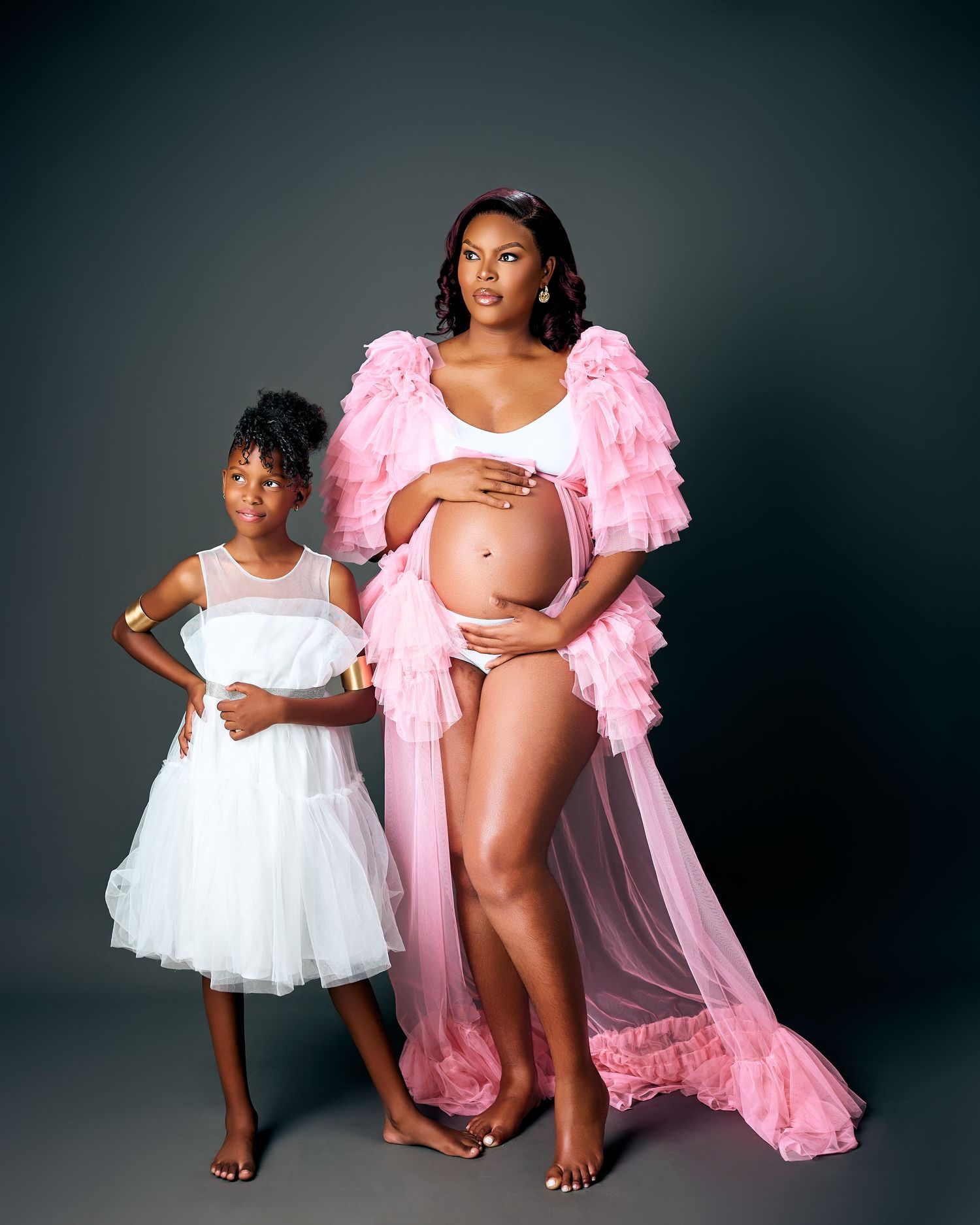 A maternity photo series showing elegant poses in pink and white flowing dresses against a dark studio backdrop.