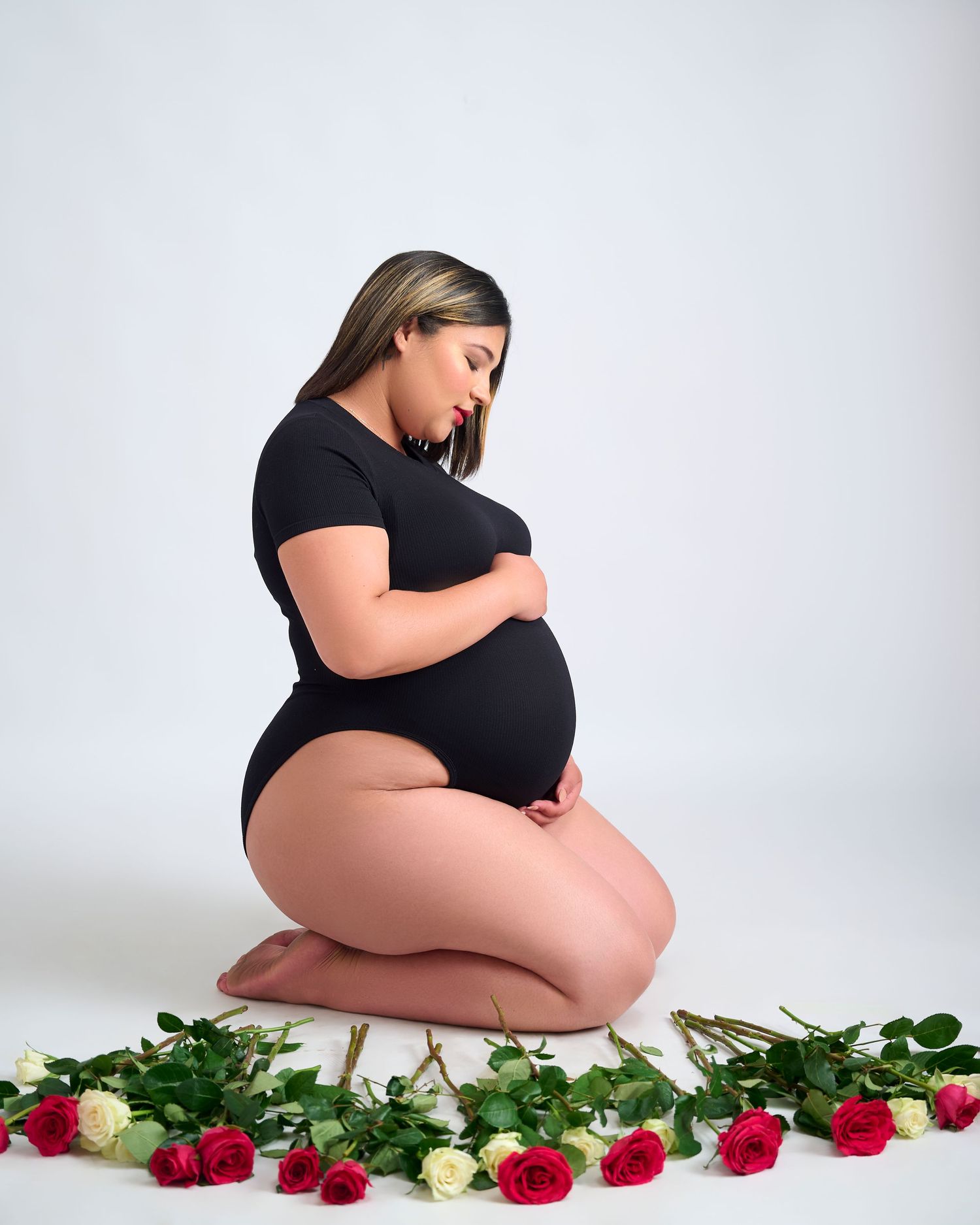 Maternity photo with a woman in black kneeling on white backdrop surrounded by red and white roses.