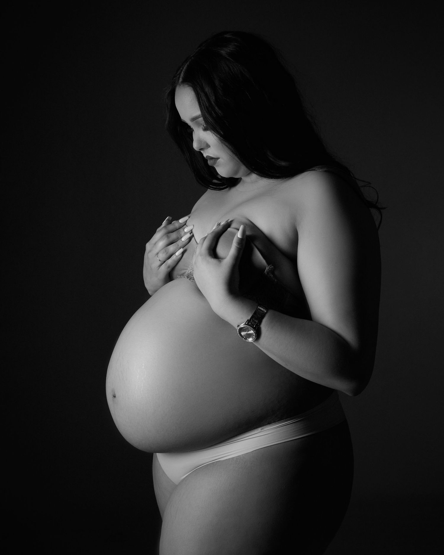 A black and white artistic maternity photo showing an expectant mother in an intimate pose against a dark background.