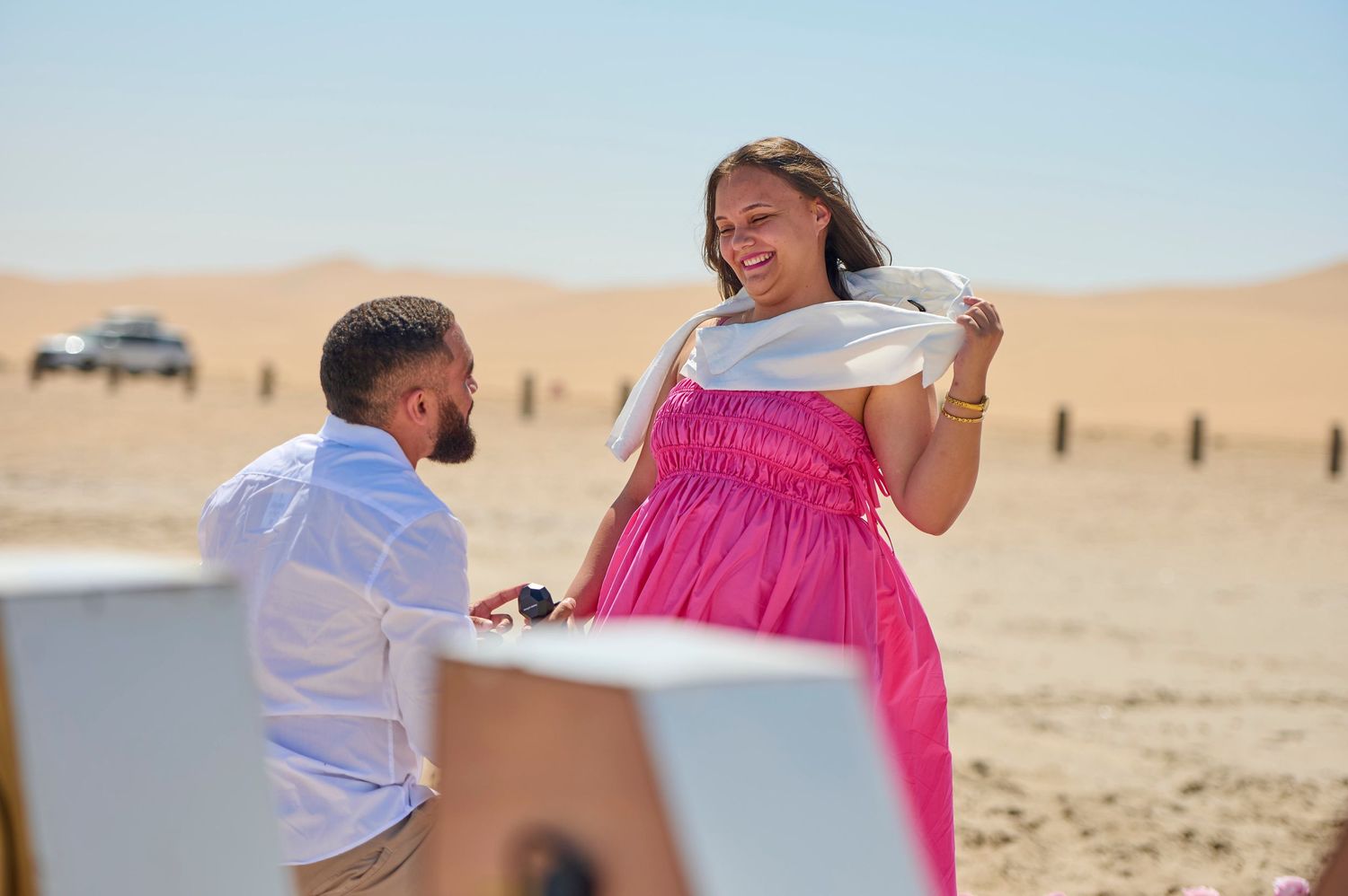 Man proposing to woman in pink dress in desert sand dunes, romantic outdoor marriage proposal scene.