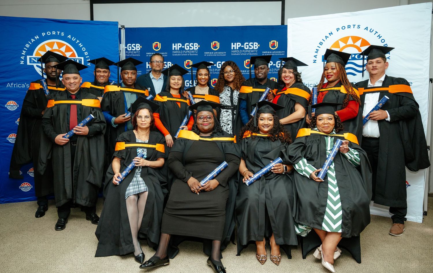 Group of graduates in black robes with orange and yellow trim pose proudly at HP-GSB university graduation ceremony.